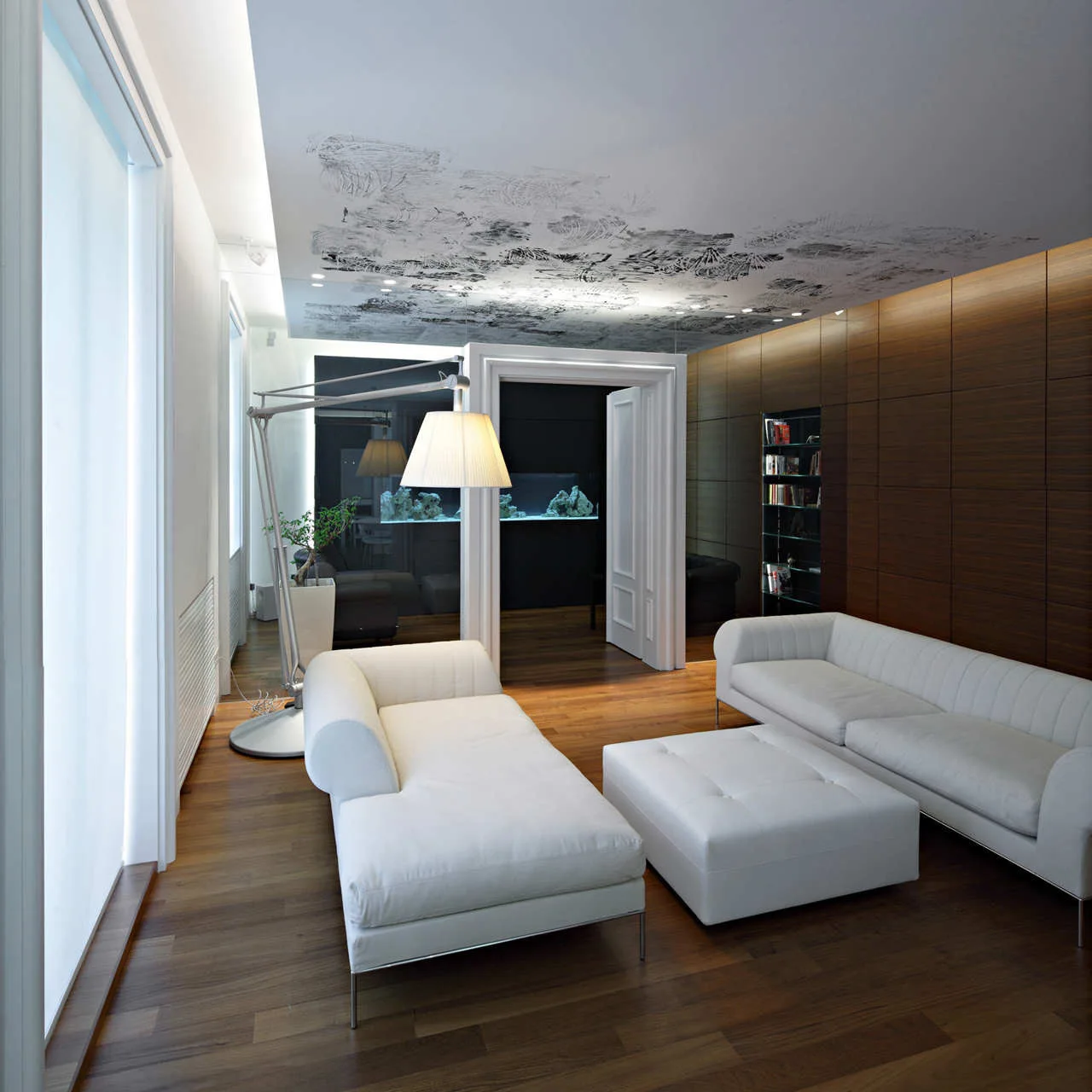 Modern living room featuring two white sofas and a matching ottoman on dark hardwood floors, with wood paneled walls and a patterned ceiling mural.