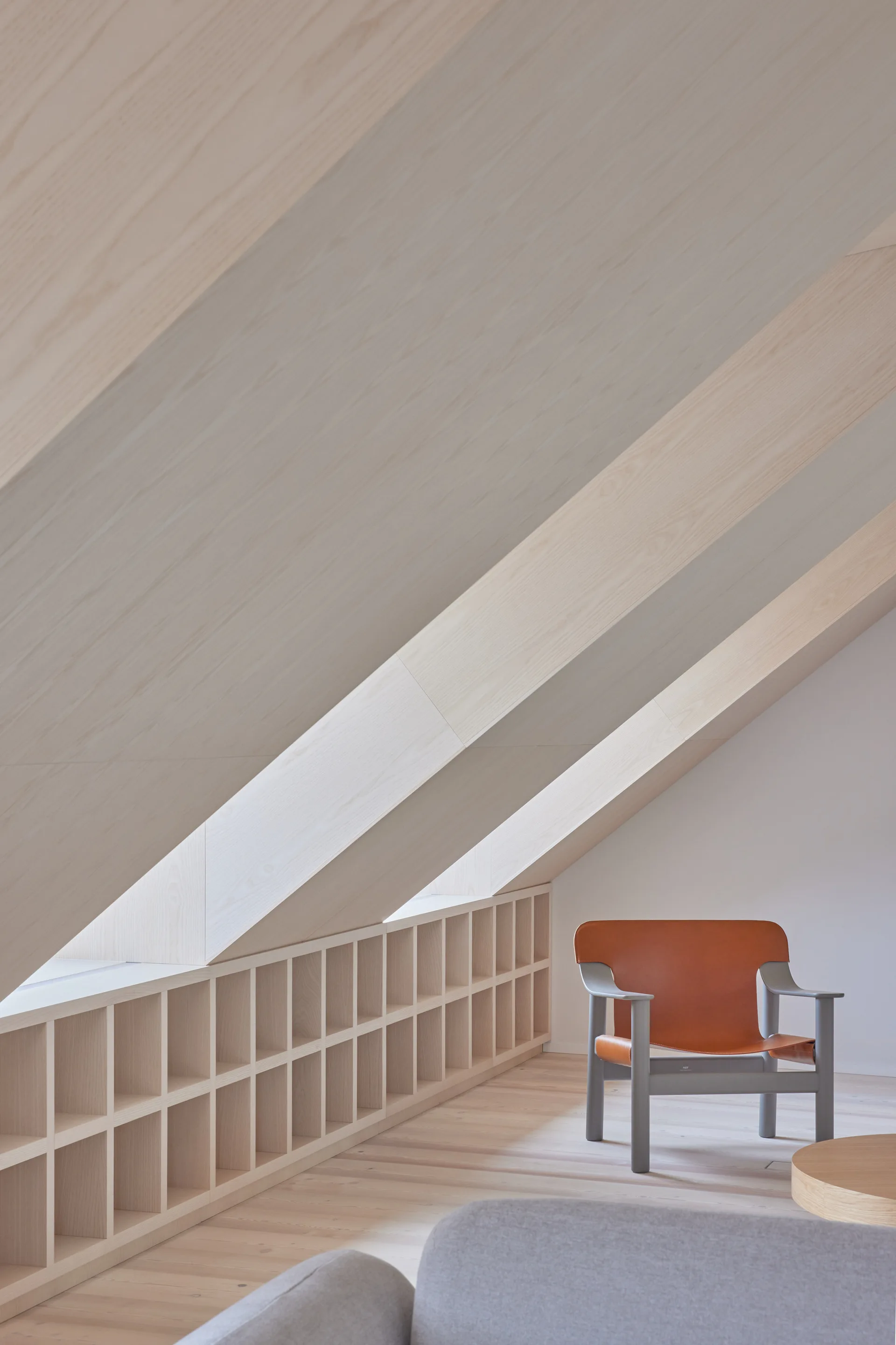 A minimalist attic living space features a sloped light wood ceiling, a low-profile grid bookshelf, and a modern grey armchair with a brown leather backrest.