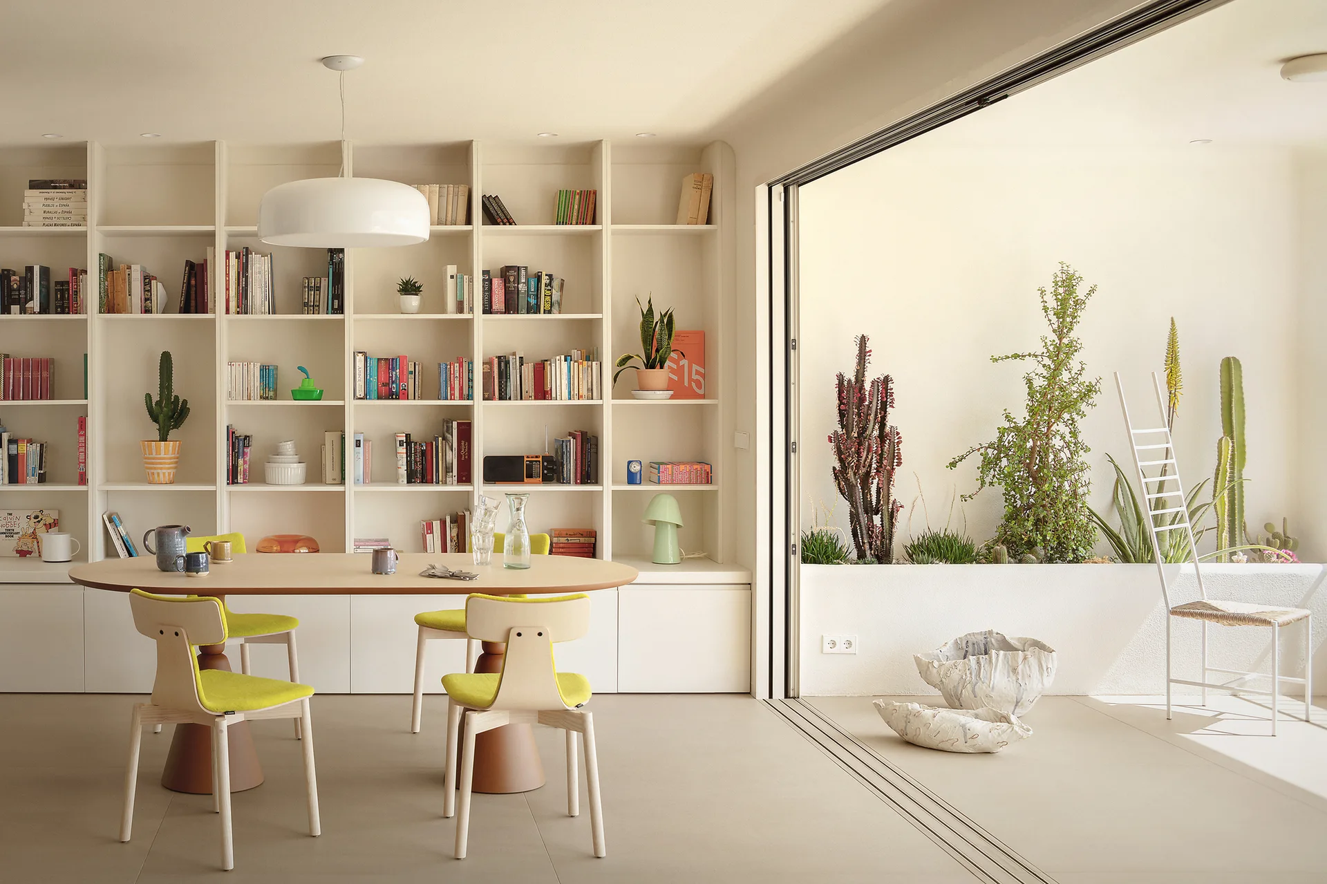 A modern dining room features an elliptical table and lime green chairs next to a large built-in bookshelf and a sliding glass door opening to a patio.