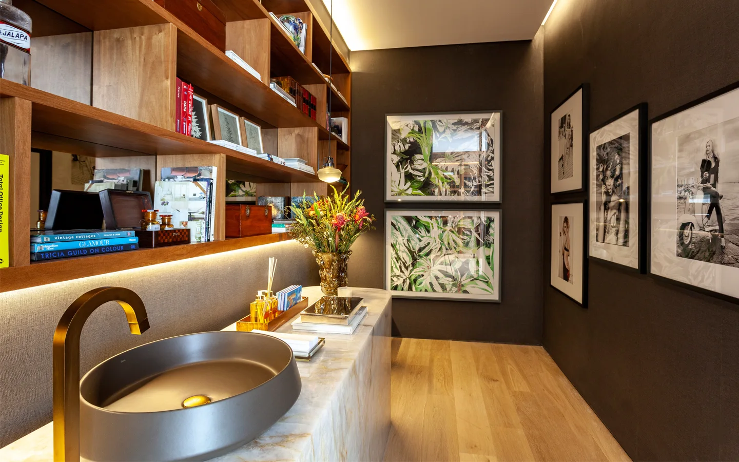 A modern powder room features a dark vessel sink on a veined stone vanity, open wooden shelving with under-cabinet lighting, and framed wall art.