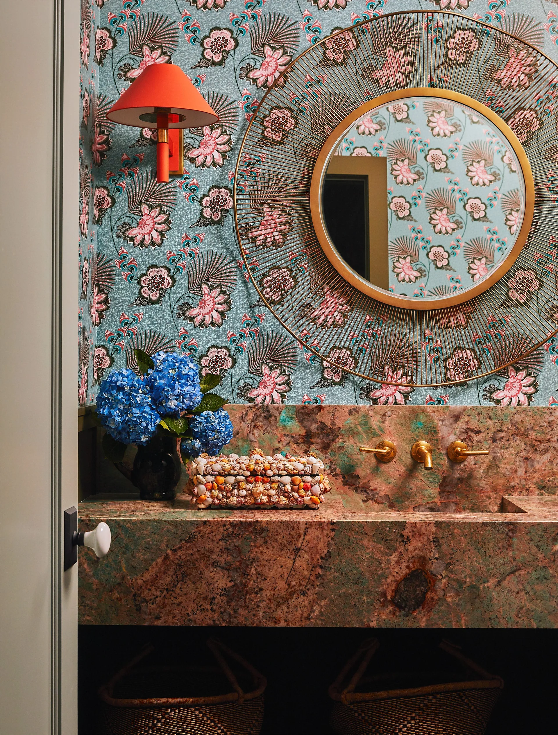 A powder room features teal floral wallpaper, a patterned pink stone vanity with integrated sink, a gold sunburst mirror, and a red wall sconce.