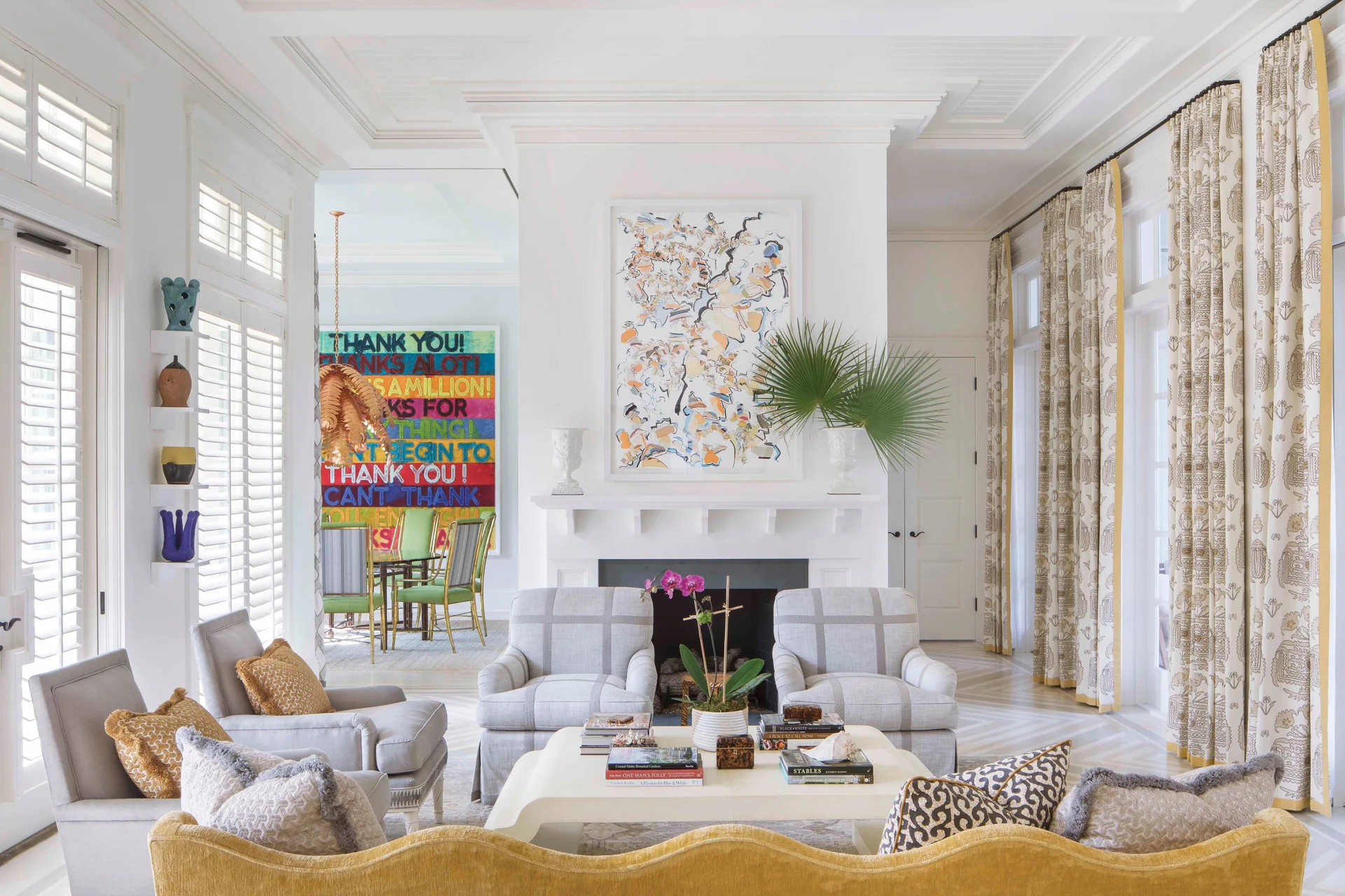A bright living room features a white fireplace, two gray plaid armchairs, a yellow sofa in the foreground, and large shuttered windows.