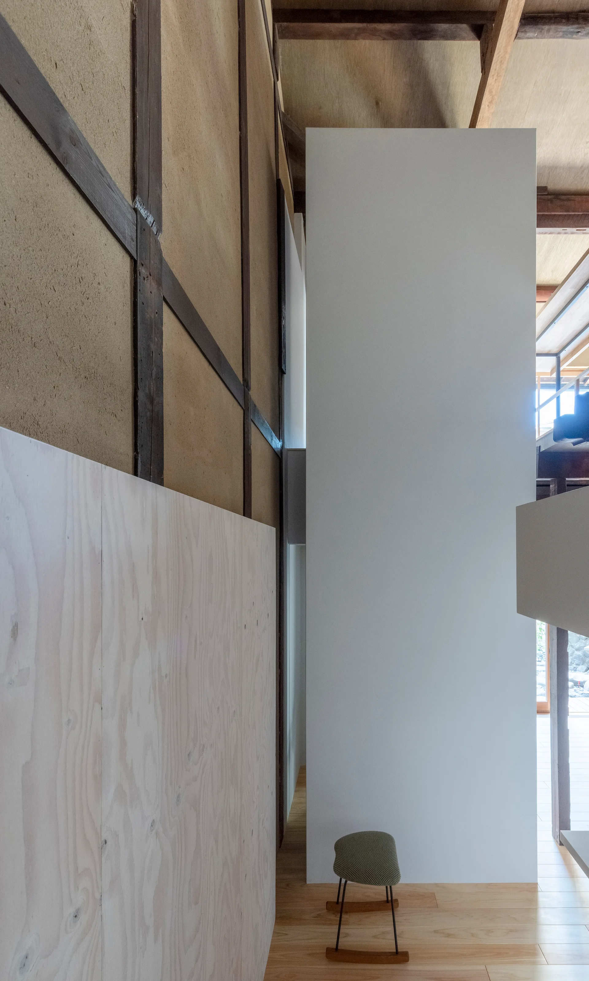 A modern minimalist interior displays a stark white structural column standing next to a traditional textured earth wall and light wood paneling.