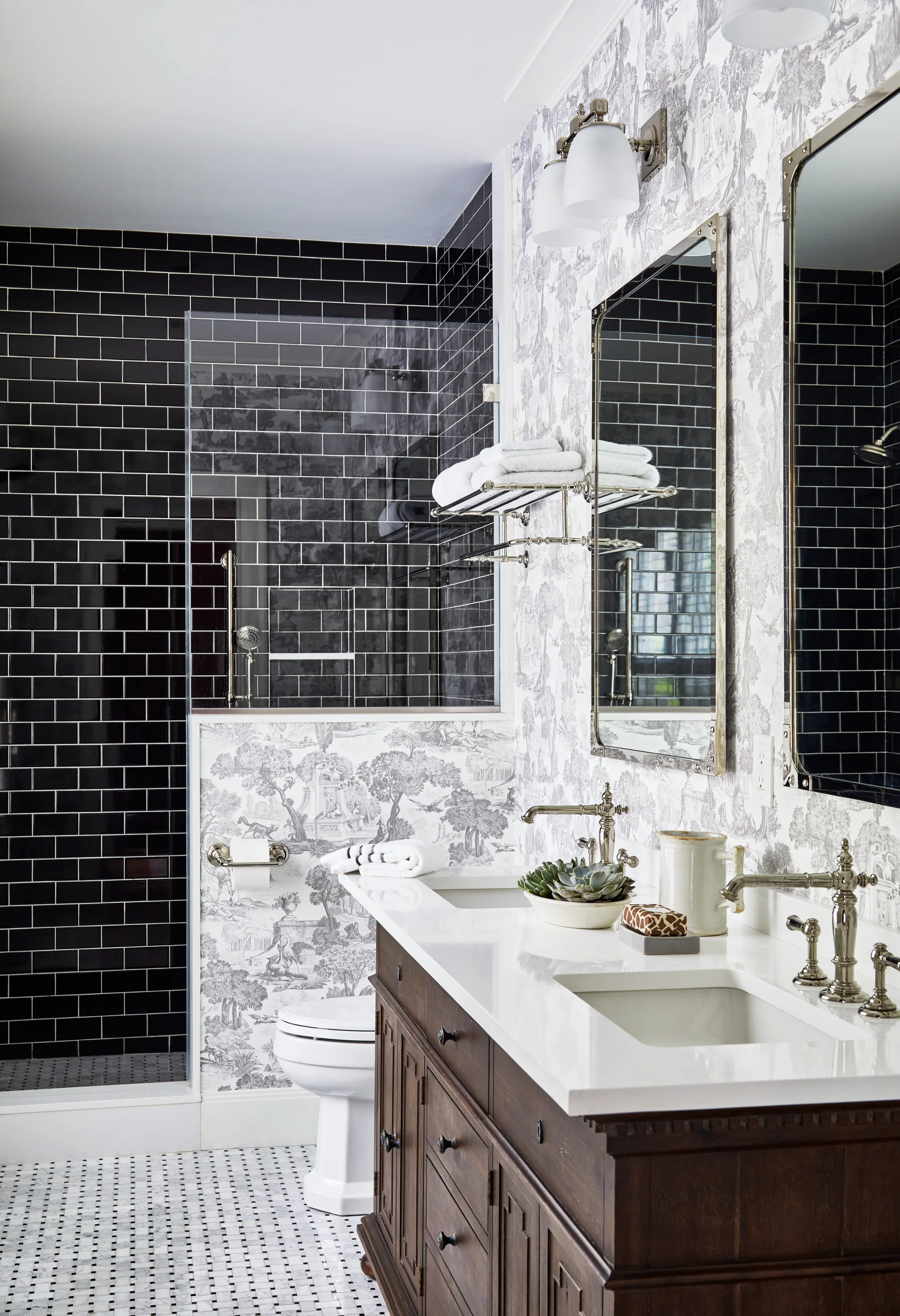 A traditional bathroom features a dark wood double vanity, black-and-white toile wallpaper, and a glass-enclosed shower lined with black subway tile.