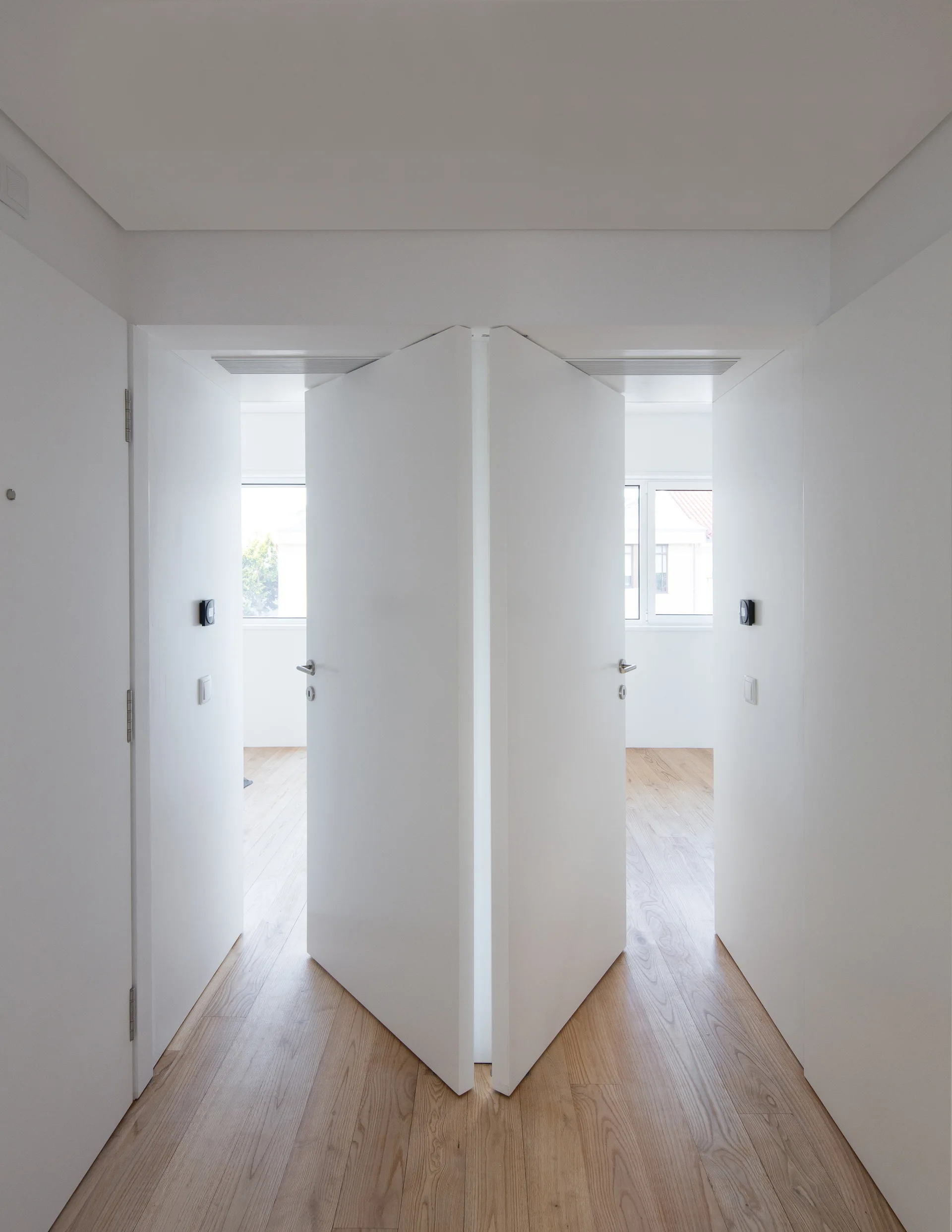 Symmetrical white doors open outward from a minimalist hallway with light wood plank flooring and white walls.