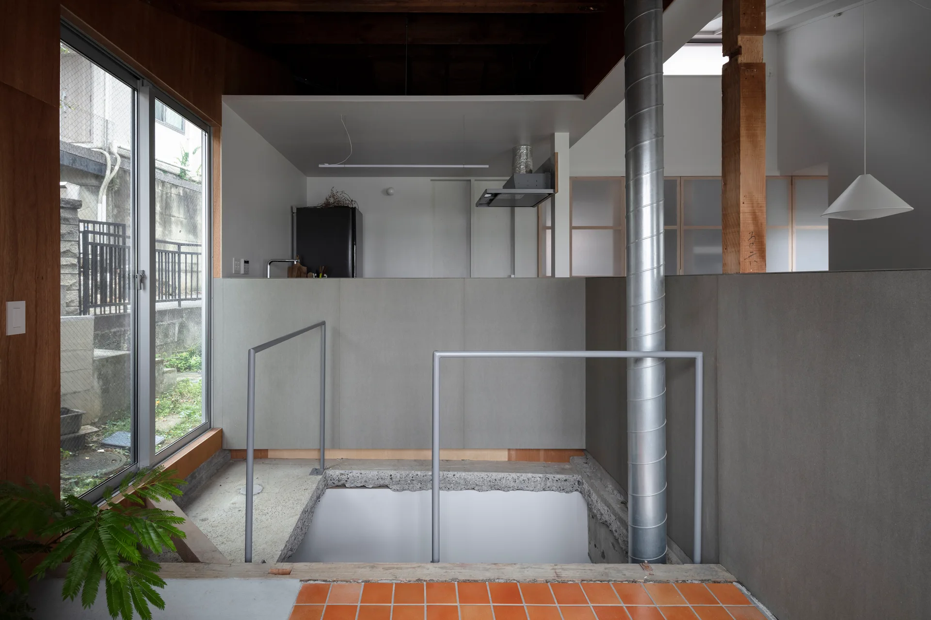 A split-level interior features a kitchen behind a grey partition, terracotta floor tiles, a vertical metal ventilation pipe, and a sunken stairwell.