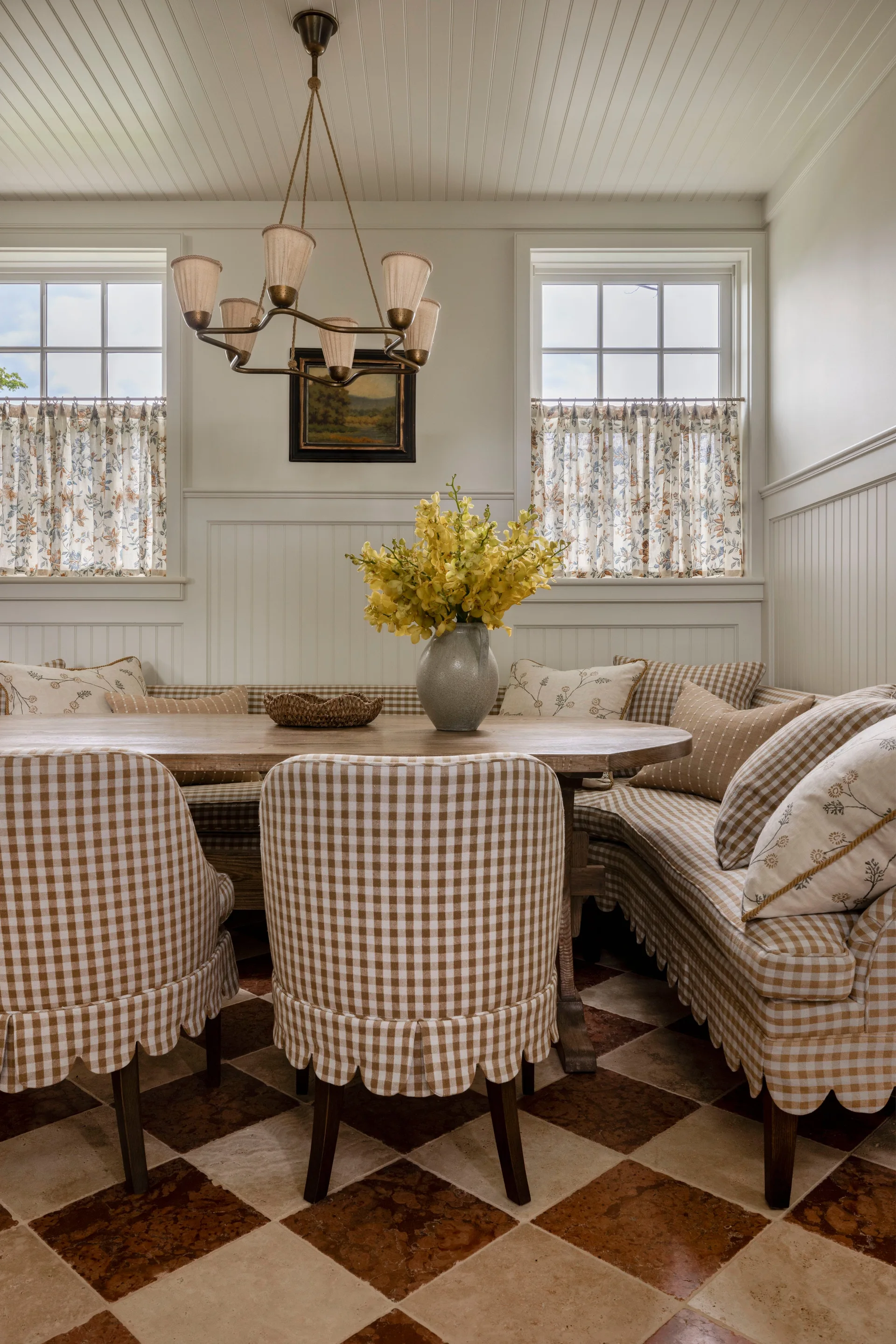 A breakfast nook features a wooden table surrounded by gingham-patterned skirted chairs and a corner banquette over a brown and beige checkered floor.