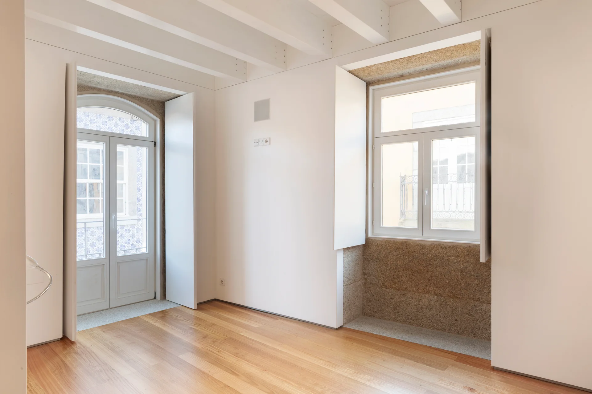 An empty room features light wood floors, white painted ceiling beams, and deep recessed windows framed in natural textured stone.