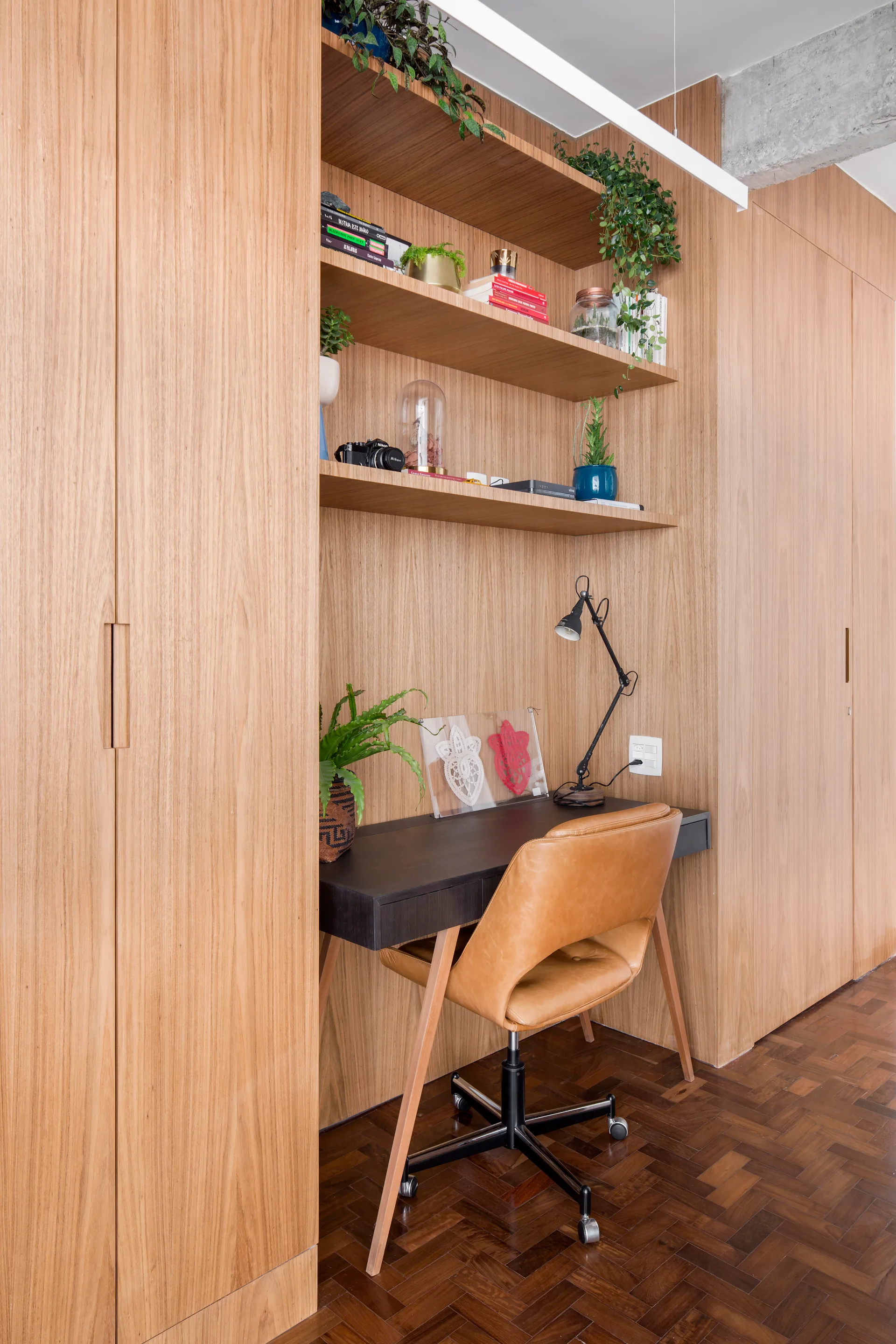 A black desk is recessed inside a large wood cabinetry wall with open shelves, paired with a tan leather chair over dark parquet floors.
