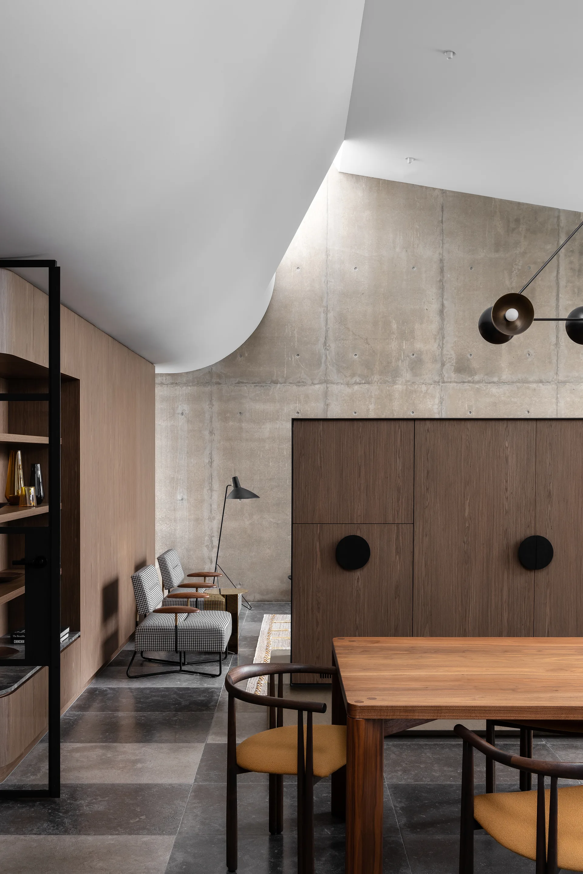 An interior dining space displays a wooden table, yellow upholstered chairs, houndstooth patterned accent chairs, and a curved ceiling against a concrete wall.