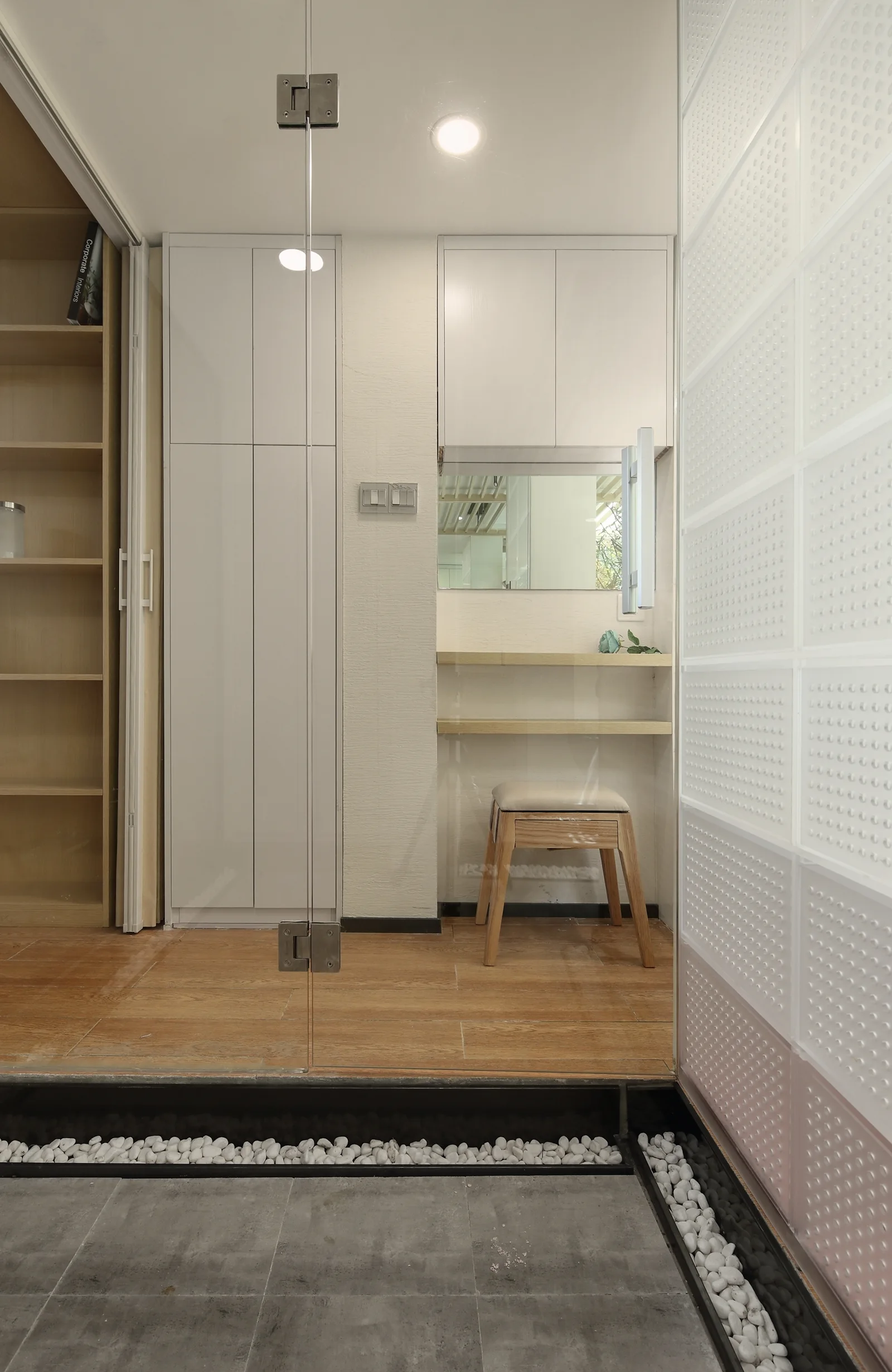A compact bathroom dressing nook features built-in white cabinets, a small wooden vanity stool, and a textured glass block partition wall.
