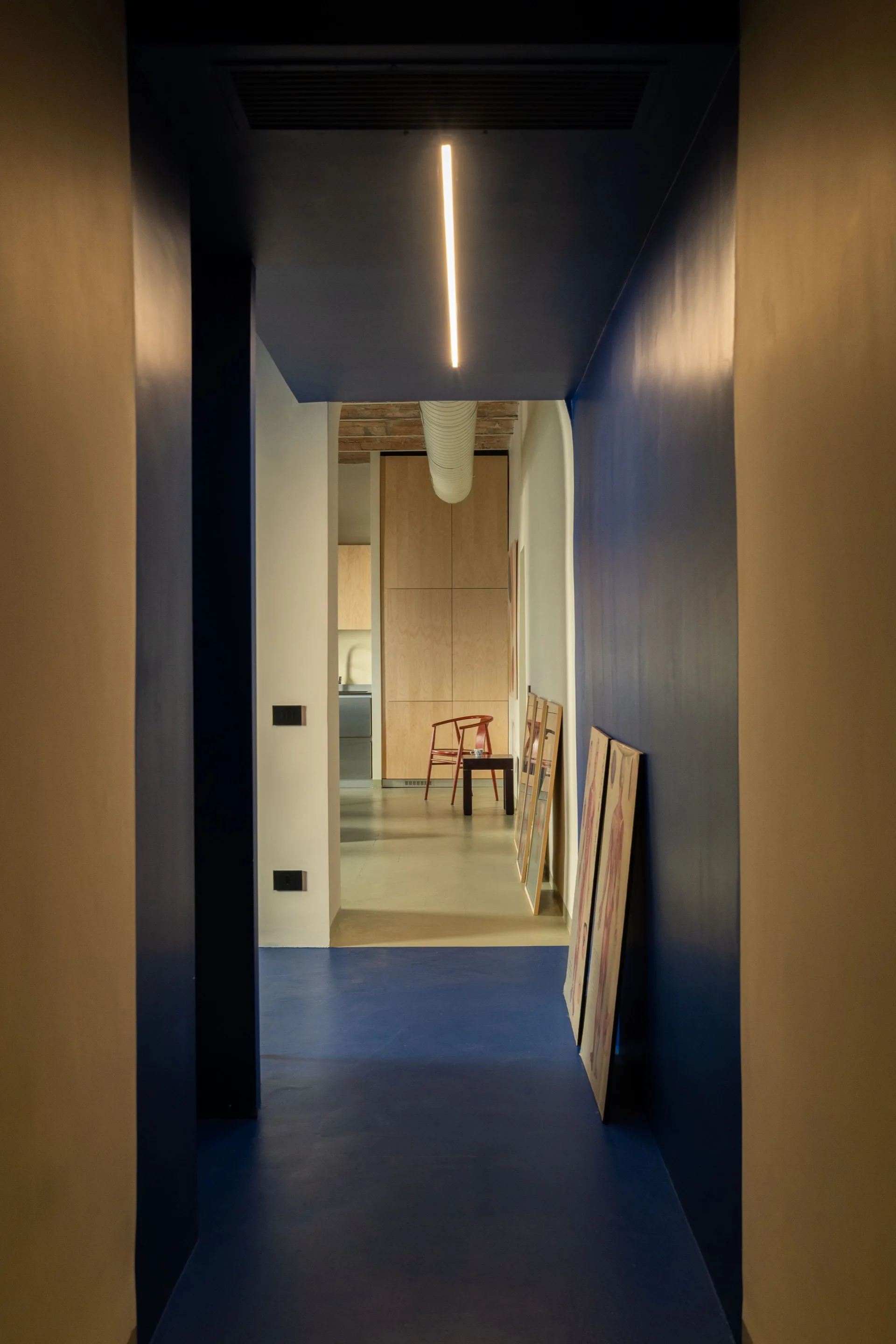 A long narrow hallway with matte blue walls and floor features a linear ceiling light and leads to a kitchen with wood-grained cabinets.