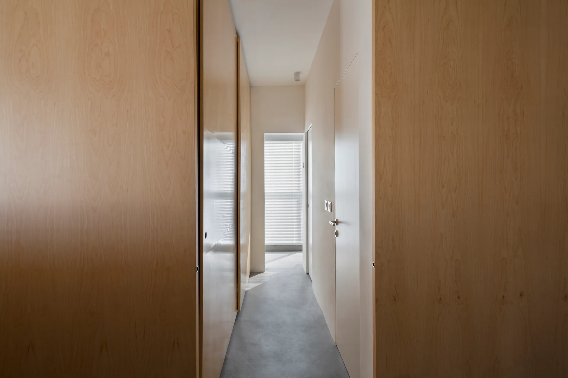 A narrow hallway featuring floor-to-ceiling light wood paneled walls, seamless flush doors, a gray concrete floor, and a window with blinds at the far end.