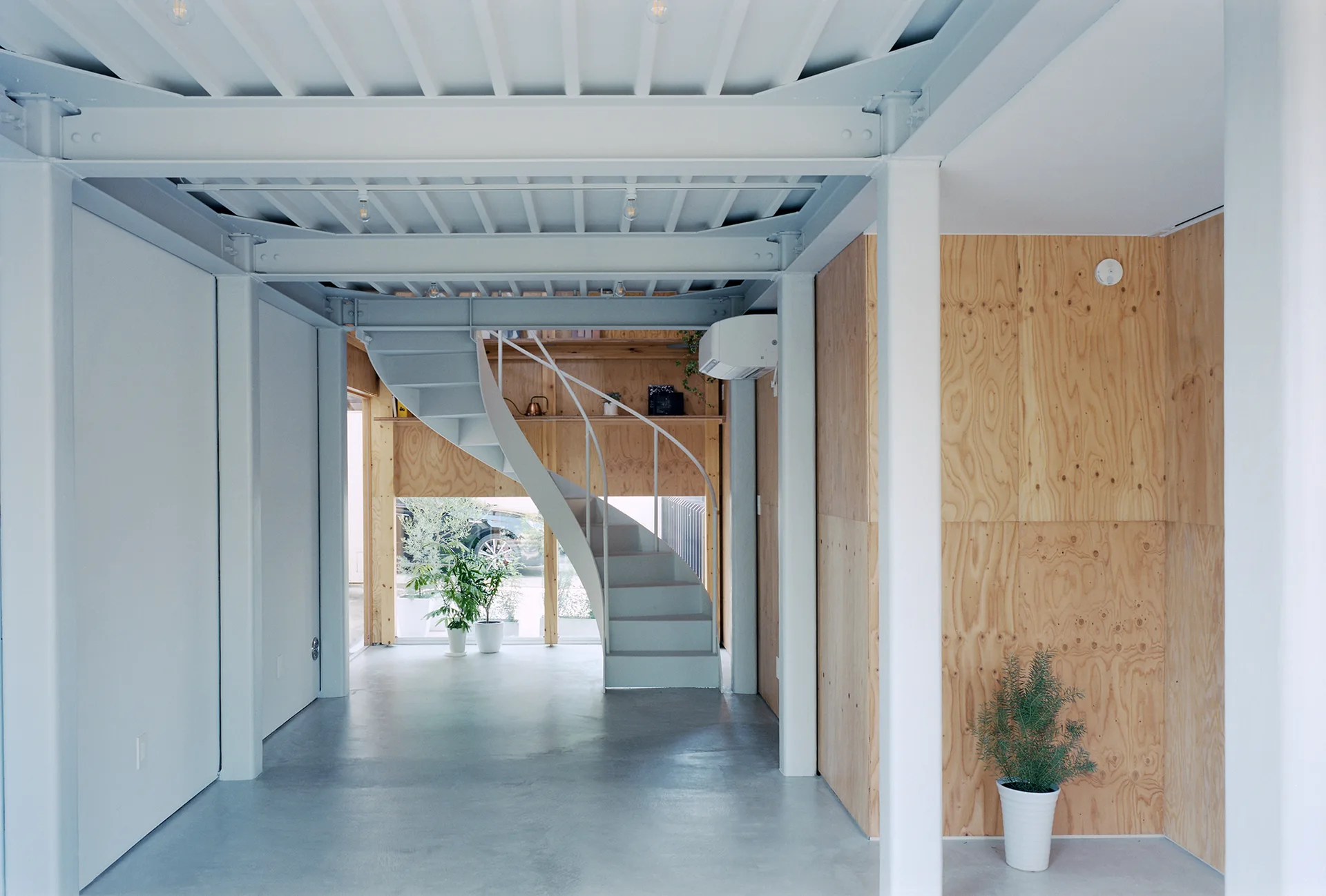 A minimalist open-plan interior featuring a white metal spiral staircase, exposed structural steel beams, and natural plywood wall panels.