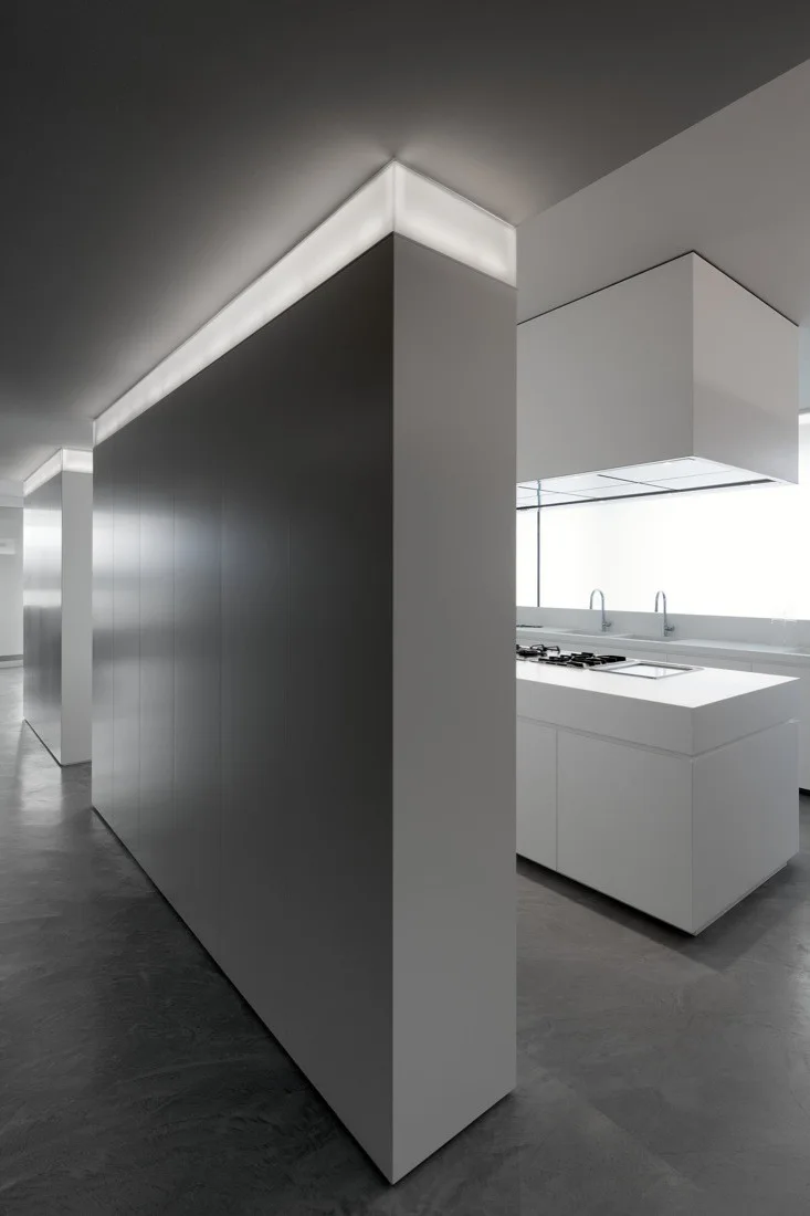 A minimalist kitchen features a monolithic grey partition wall topped with glowing cove lighting next to a solid white island.