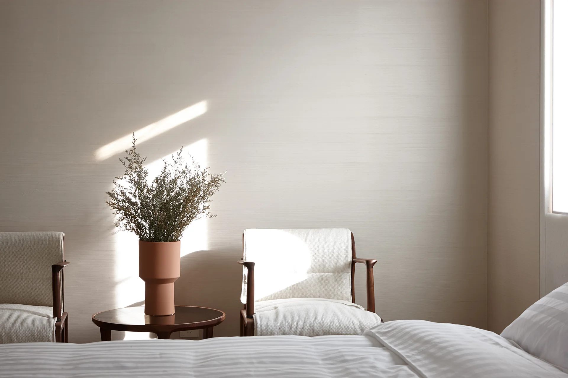 A minimalist bedroom seating area with two wooden armchairs, a small round side table, and a terracotta vase filled with dried branches.