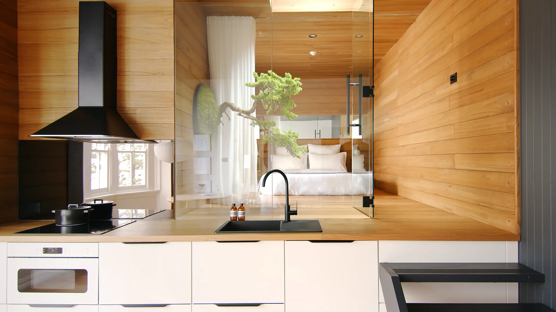 A modern kitchen with white cabinets and a black sink features a glass partition opening to a wood-paneled bedroom with a bonsai branch.