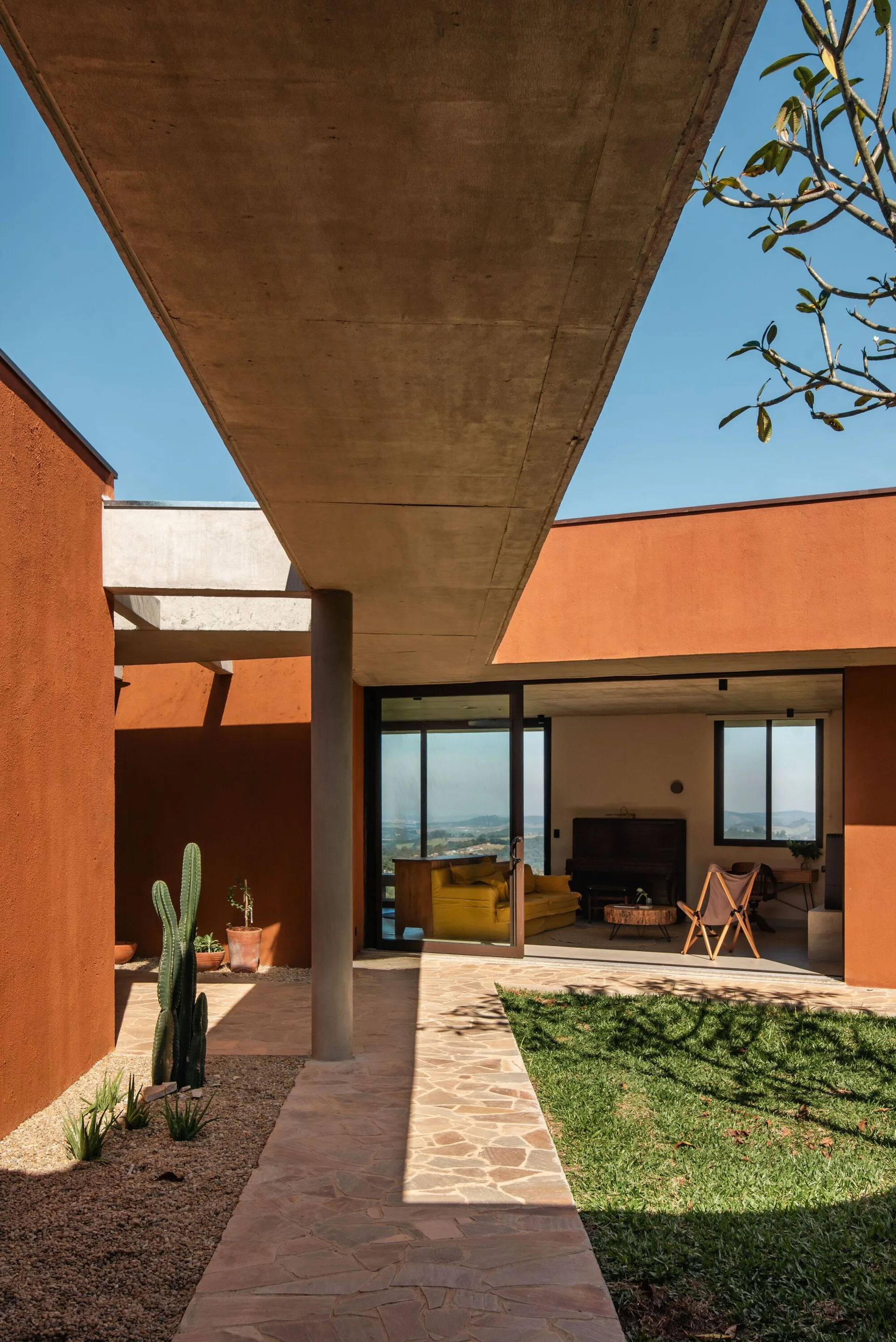 A flagstone path runs beneath a concrete overhang alongside rust-orange walls, leading to an open living room with a yellow sofa.