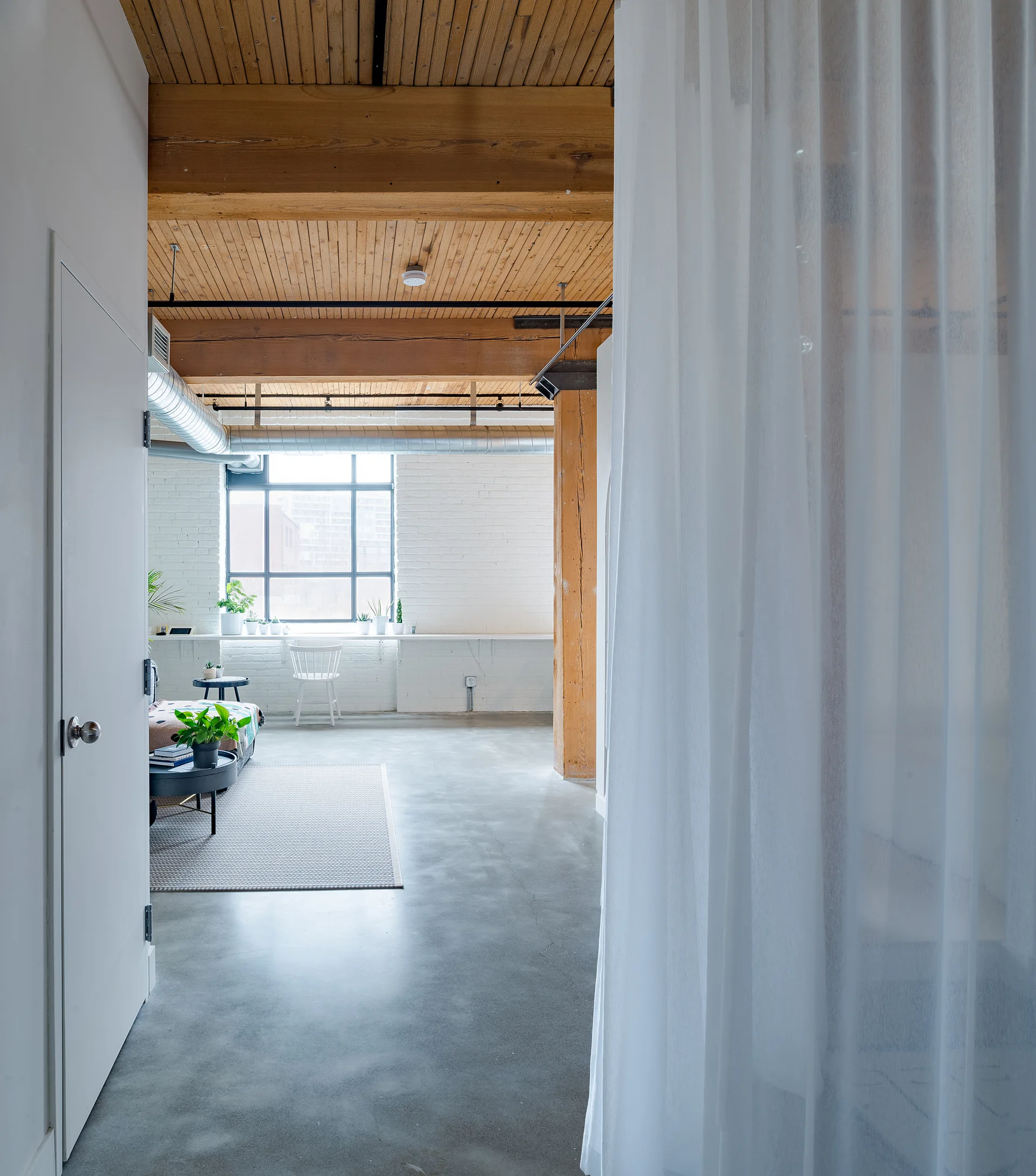 An industrial loft space featuring exposed wood ceiling beams, a white brick wall, concrete flooring, and a sheer curtain in the foreground.