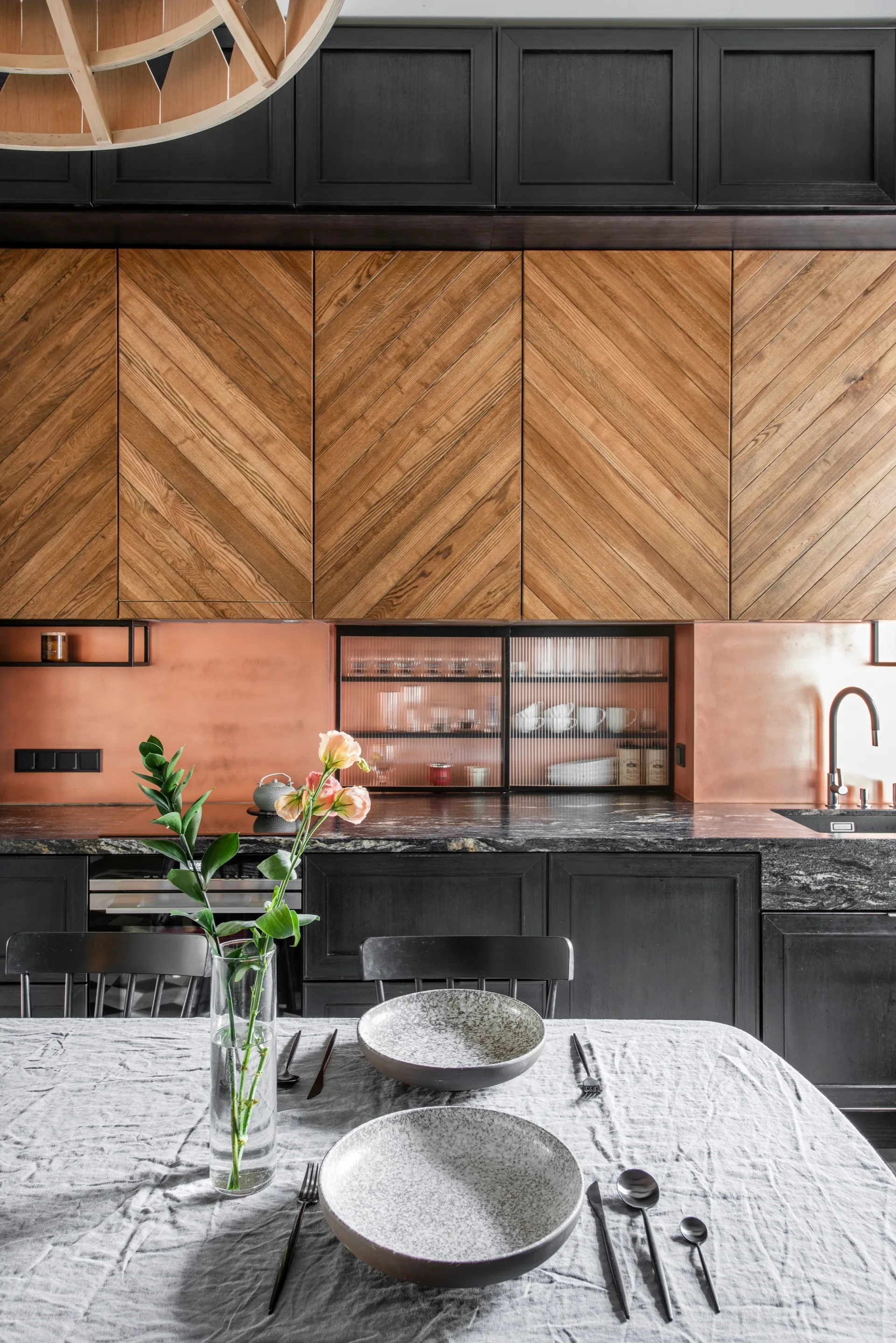 A modern kitchen features black lower cabinets, chevron-patterned wood uppers, a copper backsplash, and a dining table set in the foreground.