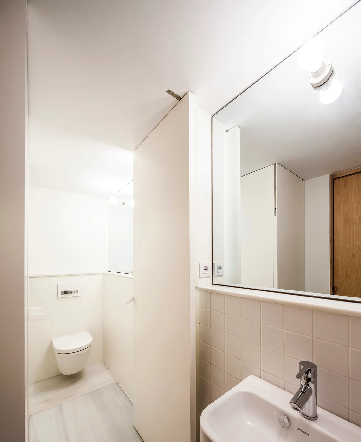 A bright white minimalist bathroom with a small sink, a large mirror over grid tile, and a wall-hung toilet behind a partition wall.