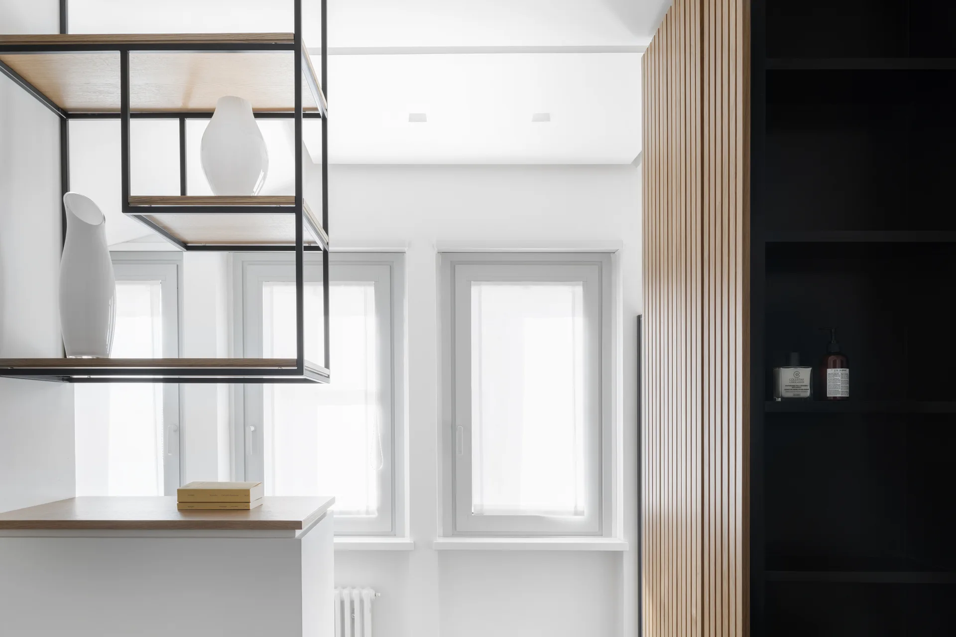 A minimalist interior showing a hanging black metal shelf with white vases next to a vertical wood slat wall and white-framed windows.