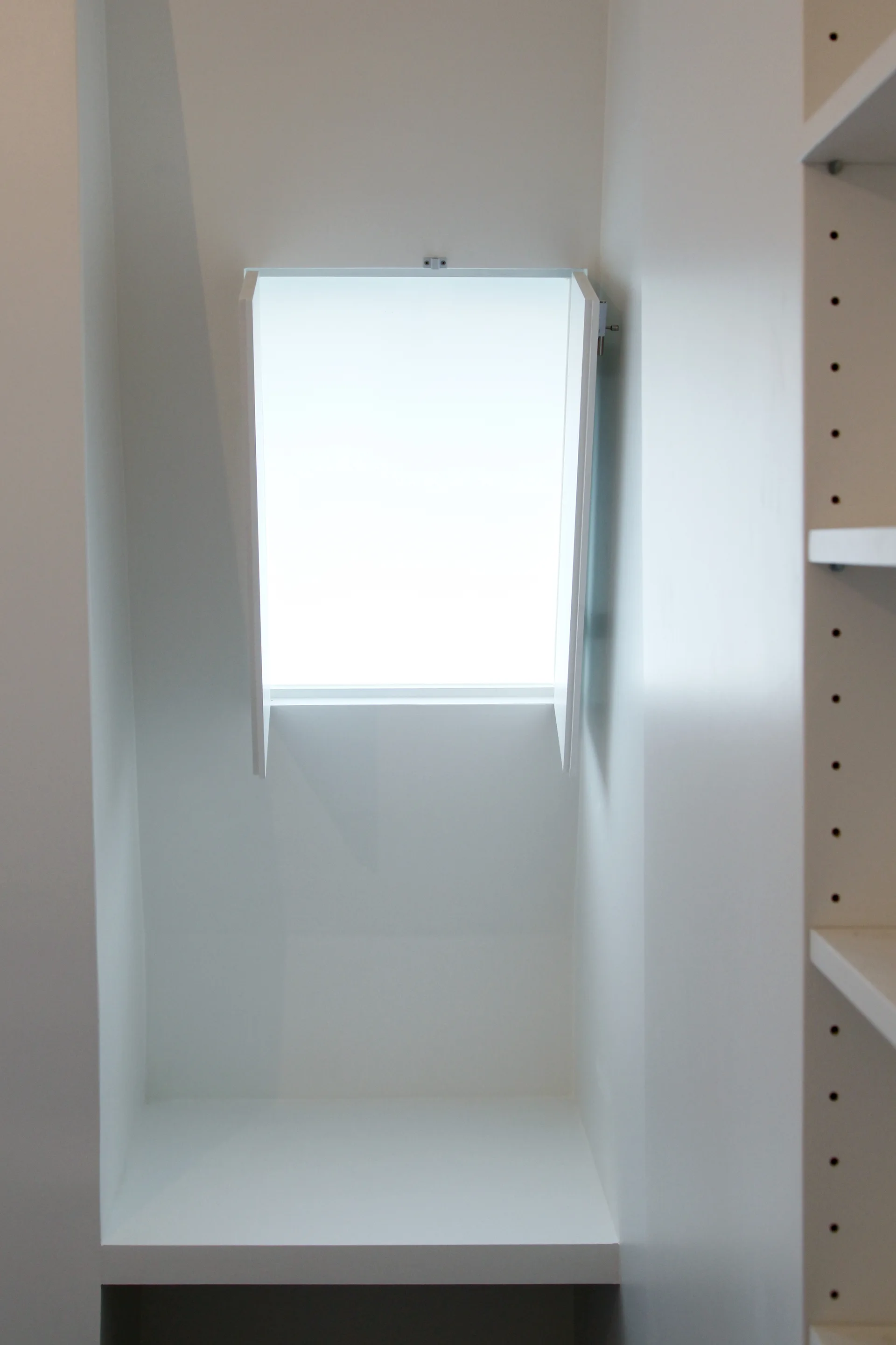 A minimalist white recessed wall niche features a small window letting in natural light, situated next to adjustable built-in shelving.