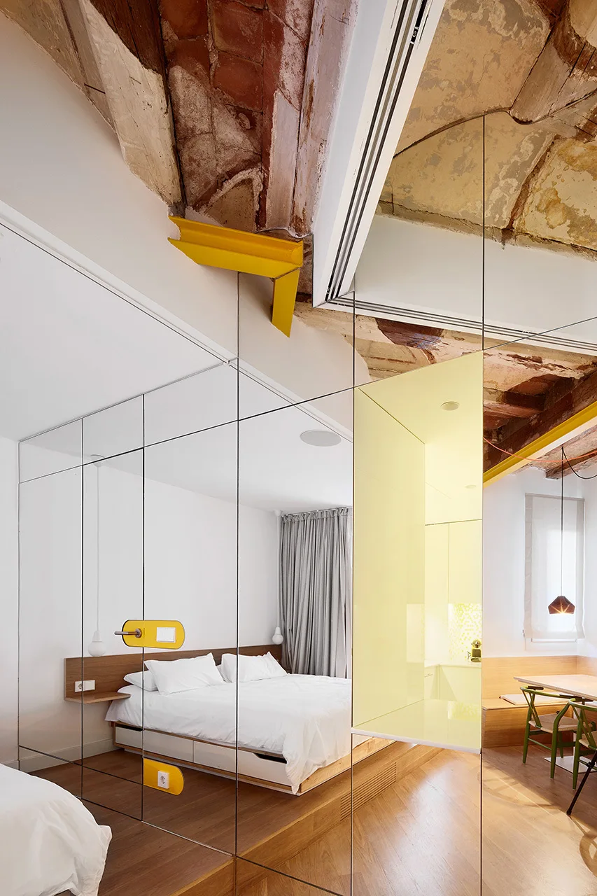 A contemporary bedroom features a platform bed set against a full-height mirrored partition wall beneath an exposed vaulted brick ceiling.