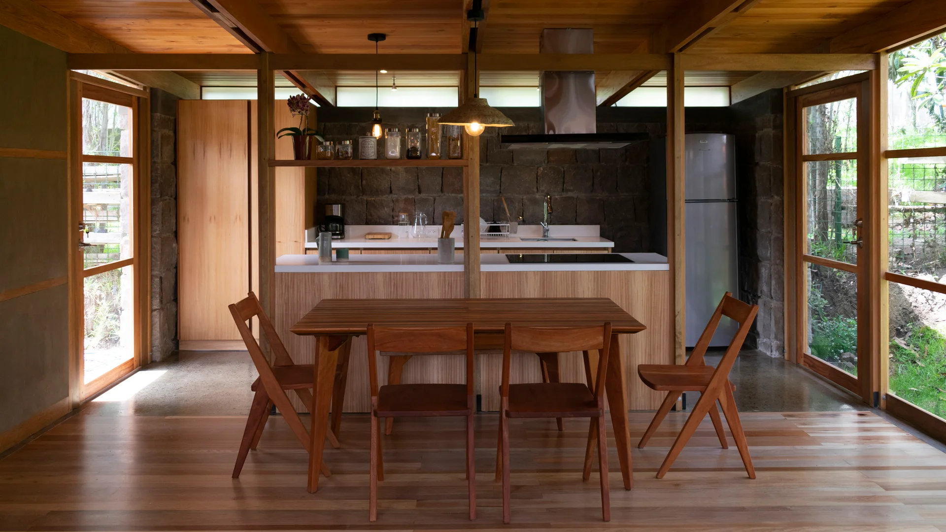 A wood-clad kitchen features a central island, a matching dining table with chairs, a textured stone backsplash wall, and large glass doors.