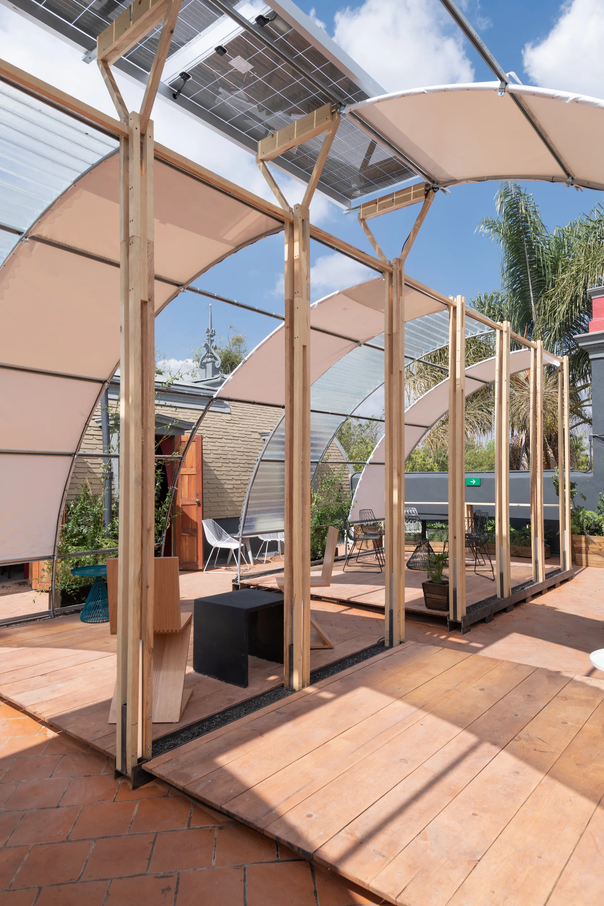An outdoor rooftop terrace features a large arched timber pergola with white fabric panels, solar panels, and wooden decking over terracotta tiles.