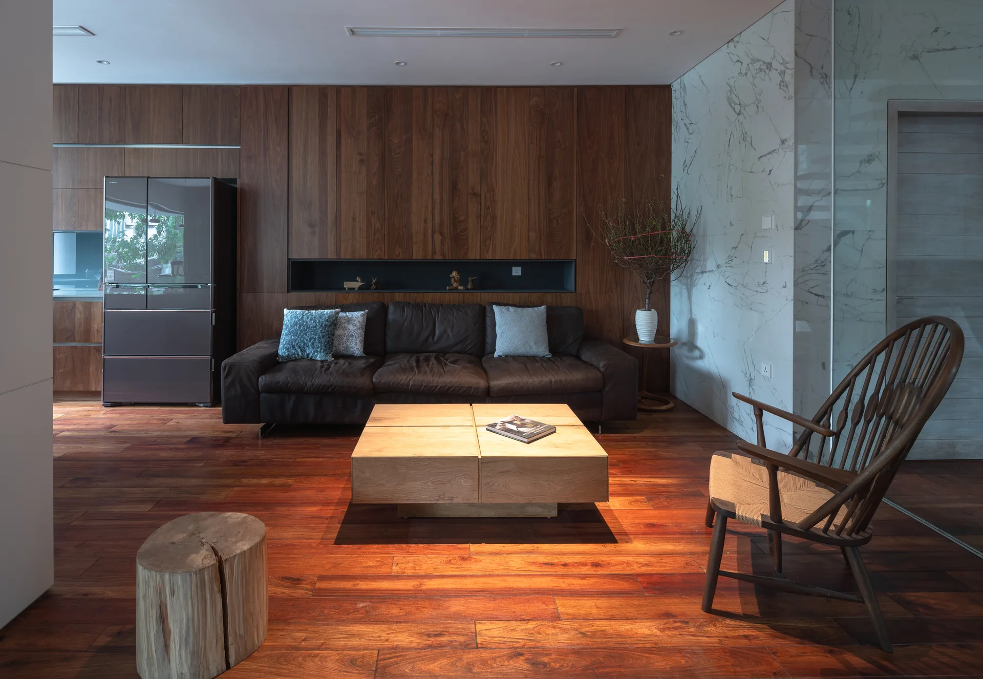A contemporary living room featuring a dark leather sofa against a warm wood paneled wall, a light oak coffee table, and a white marble accent wall.