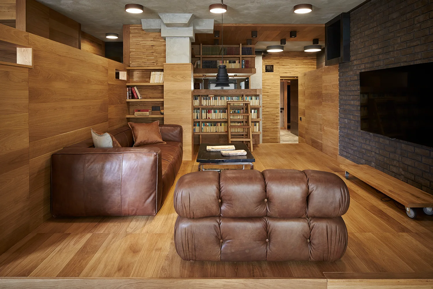 A warm wood-paneled living room features brown leather sofas, built-in bookshelves with a ladder, and a dark brick accent wall under a concrete ceiling.