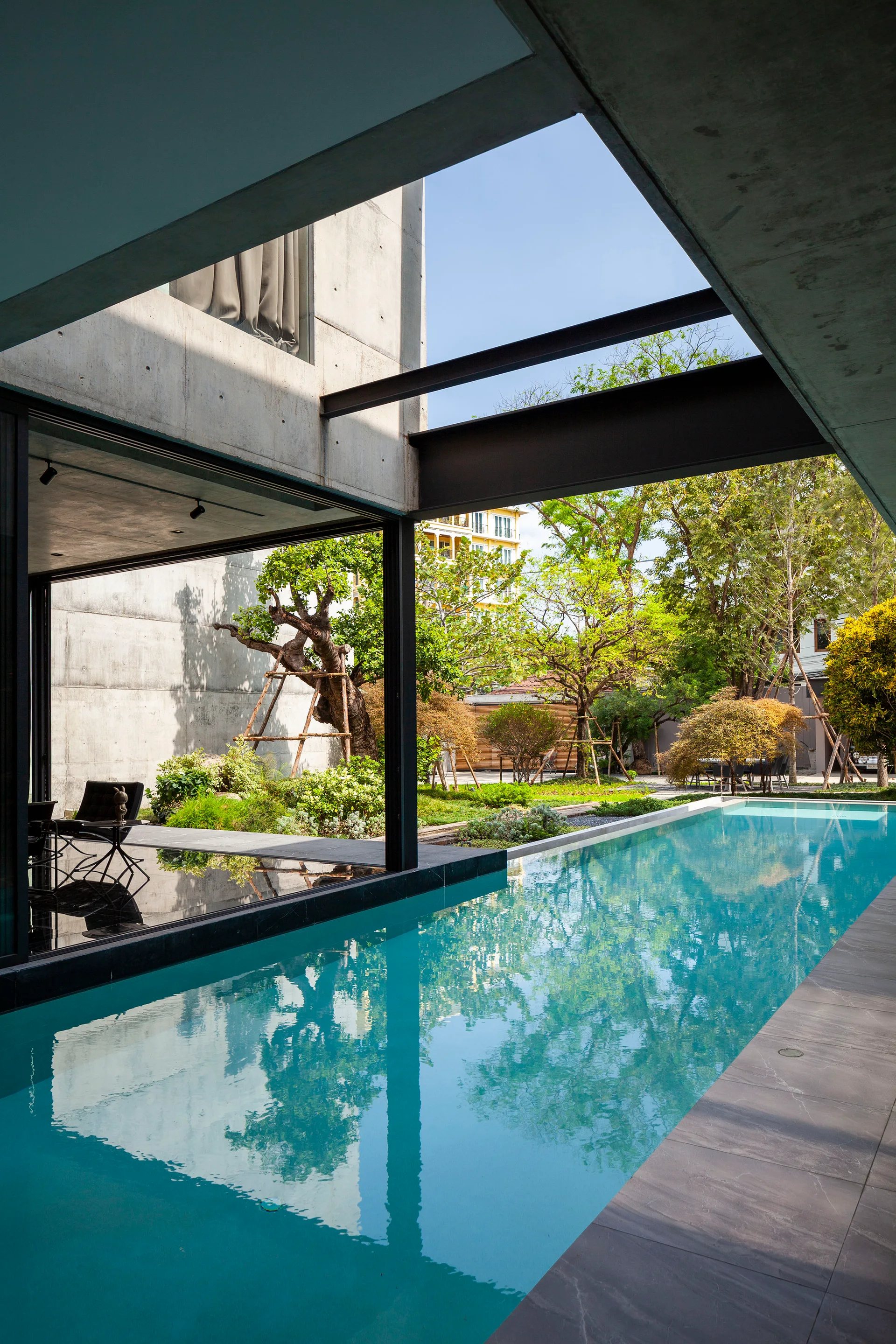 A rectangular turquoise swimming pool runs alongside an open-sided modern concrete building framed by black steel beams and lush trees.