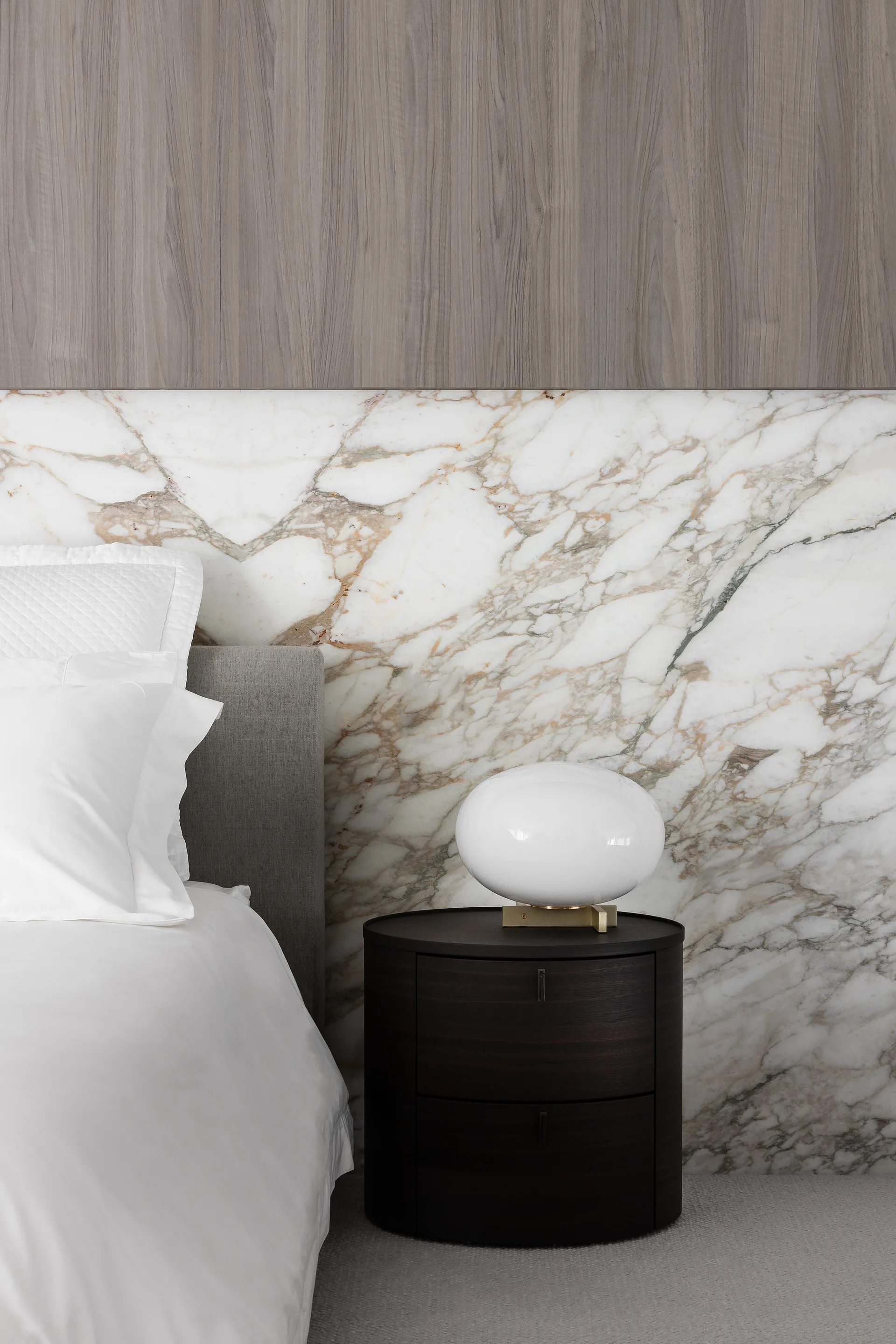 A dark wood bedside table with a white oval lamp stands next to a bed against a wall of veined white marble and grey wood paneling.