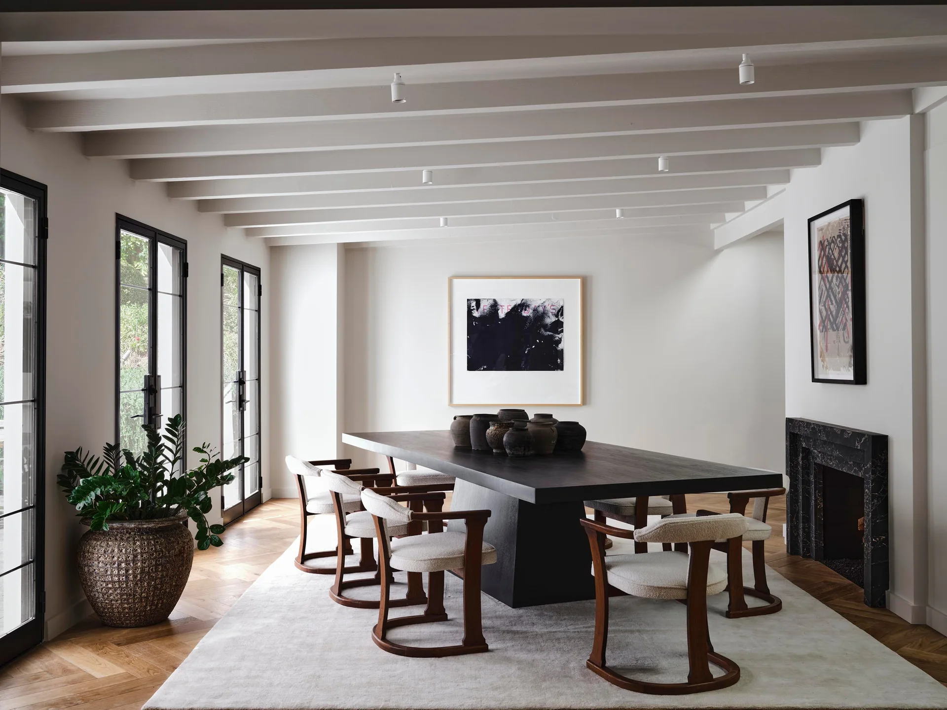 A minimalist dining room features a dark wood table with curved cream chairs over a light rug, underneath white-painted exposed ceiling beams.