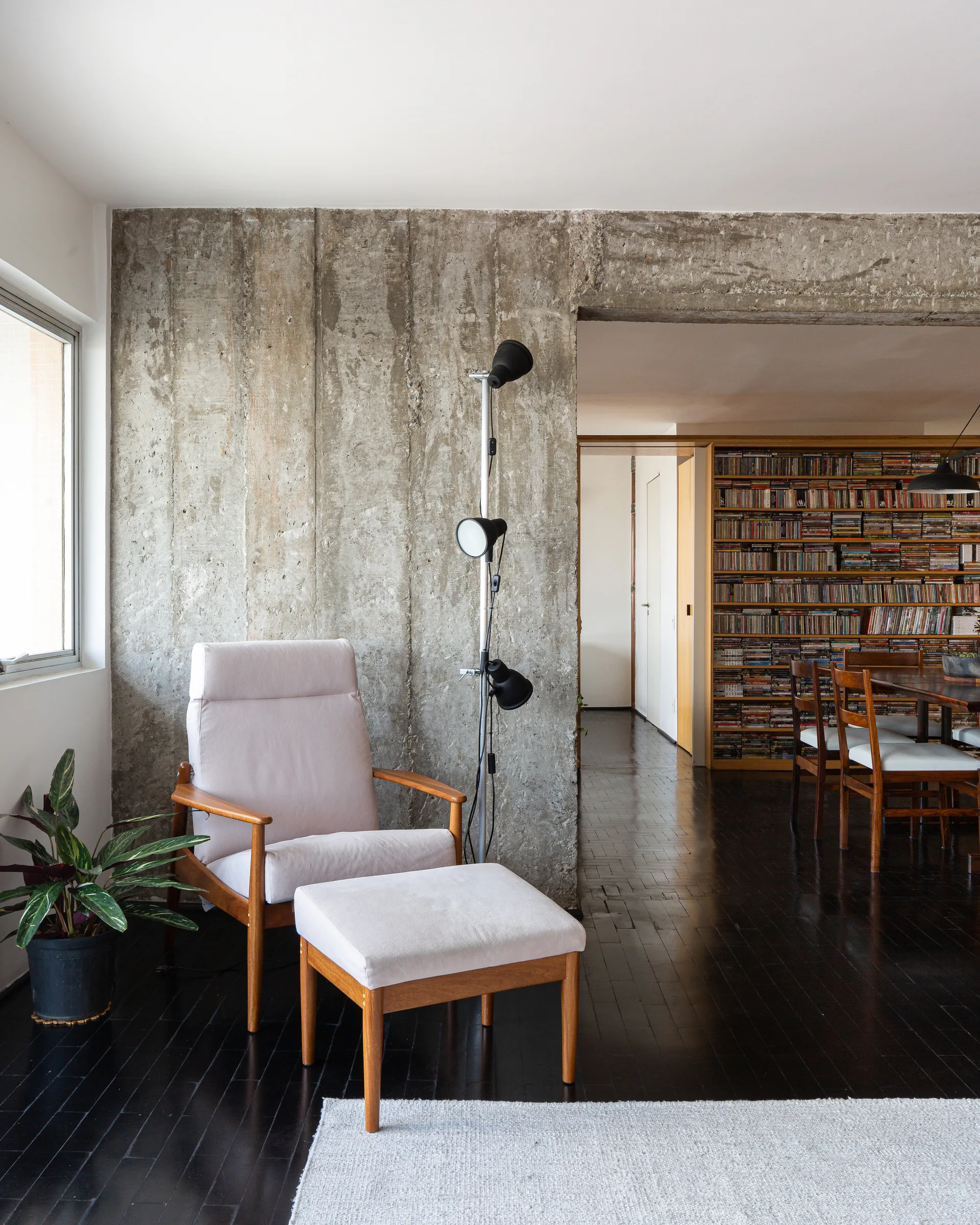 A light-colored mid-century armchair and matching ottoman sit on dark wood floors in front of a raw, textured concrete wall next to a tall black floor lamp.