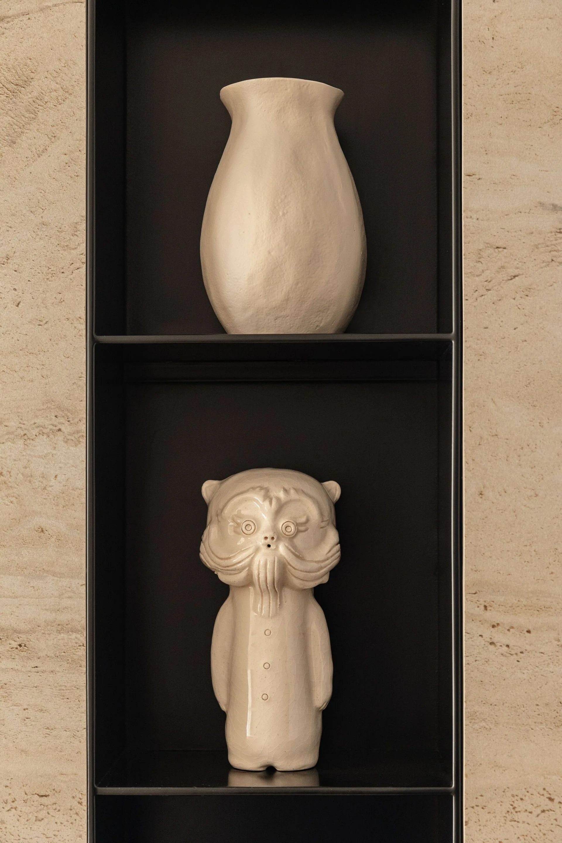 A recessed black wall niche set in a beige textured stone wall displays a matte cream ceramic vase and a whimsical glazed figurine.