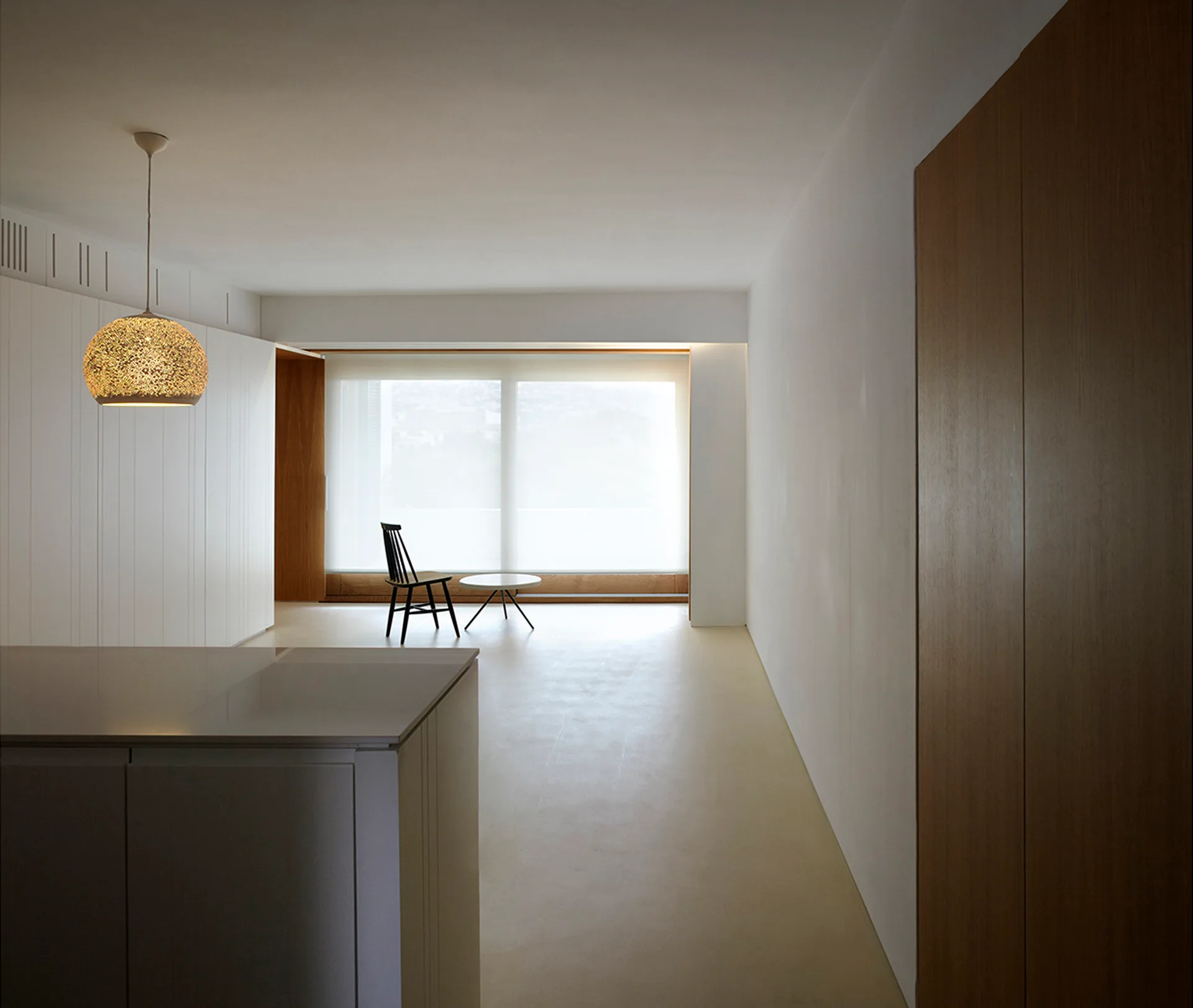 A minimalist white living space featuring built-in cabinetry, a black spindle chair, and a glowing textured pendant light.