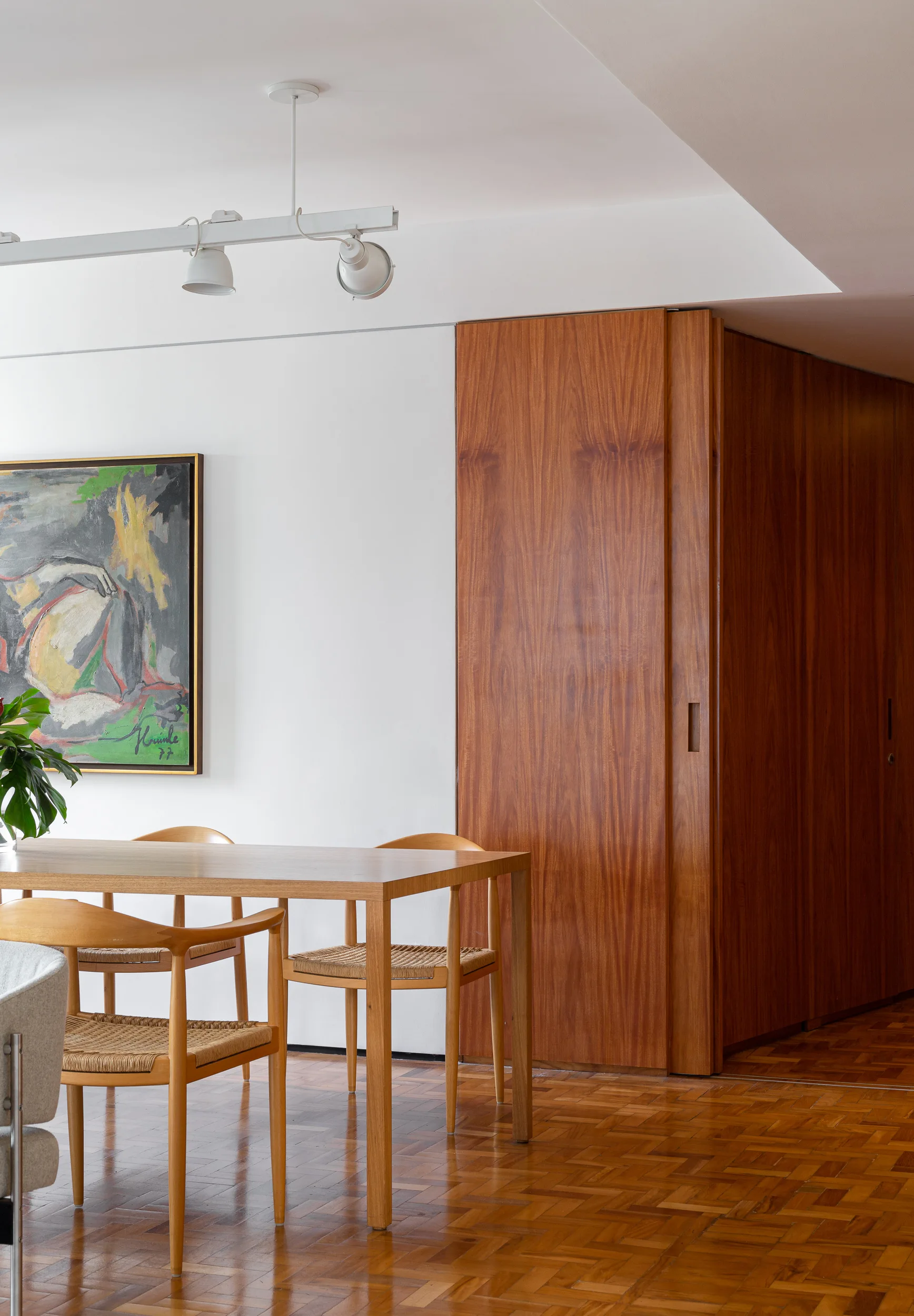 A wooden dining table and chairs with woven seats stand on parquet flooring next to a full-height wood paneled wall and white painted wall.