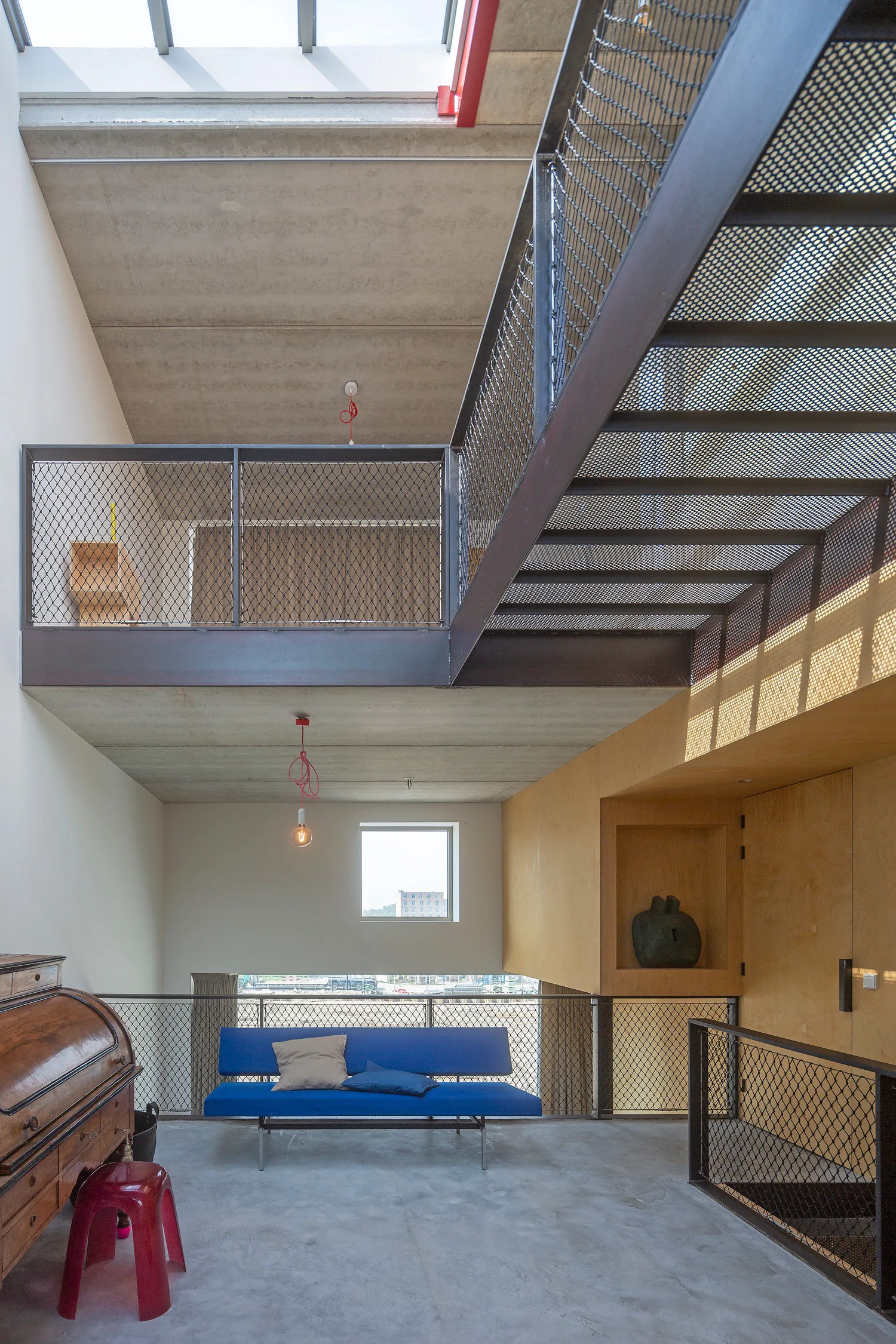 A contemporary industrial mezzanine features a bold blue sofa, concrete ceiling, warm plywood alcove, and a steel mesh staircase.