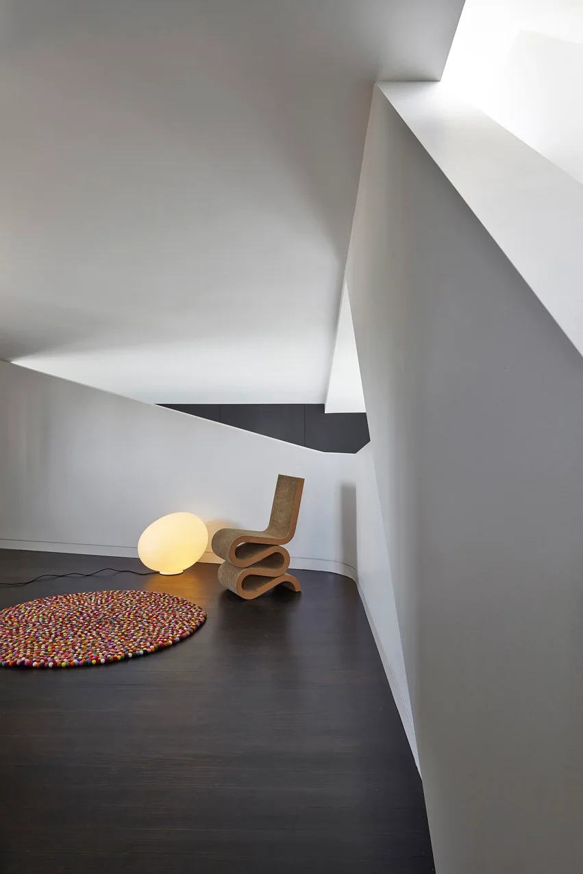 A minimalist staircase landing features a sculptural wavy chair, a glowing oval floor lamp, and a multicolored circular rug on dark wood floors.