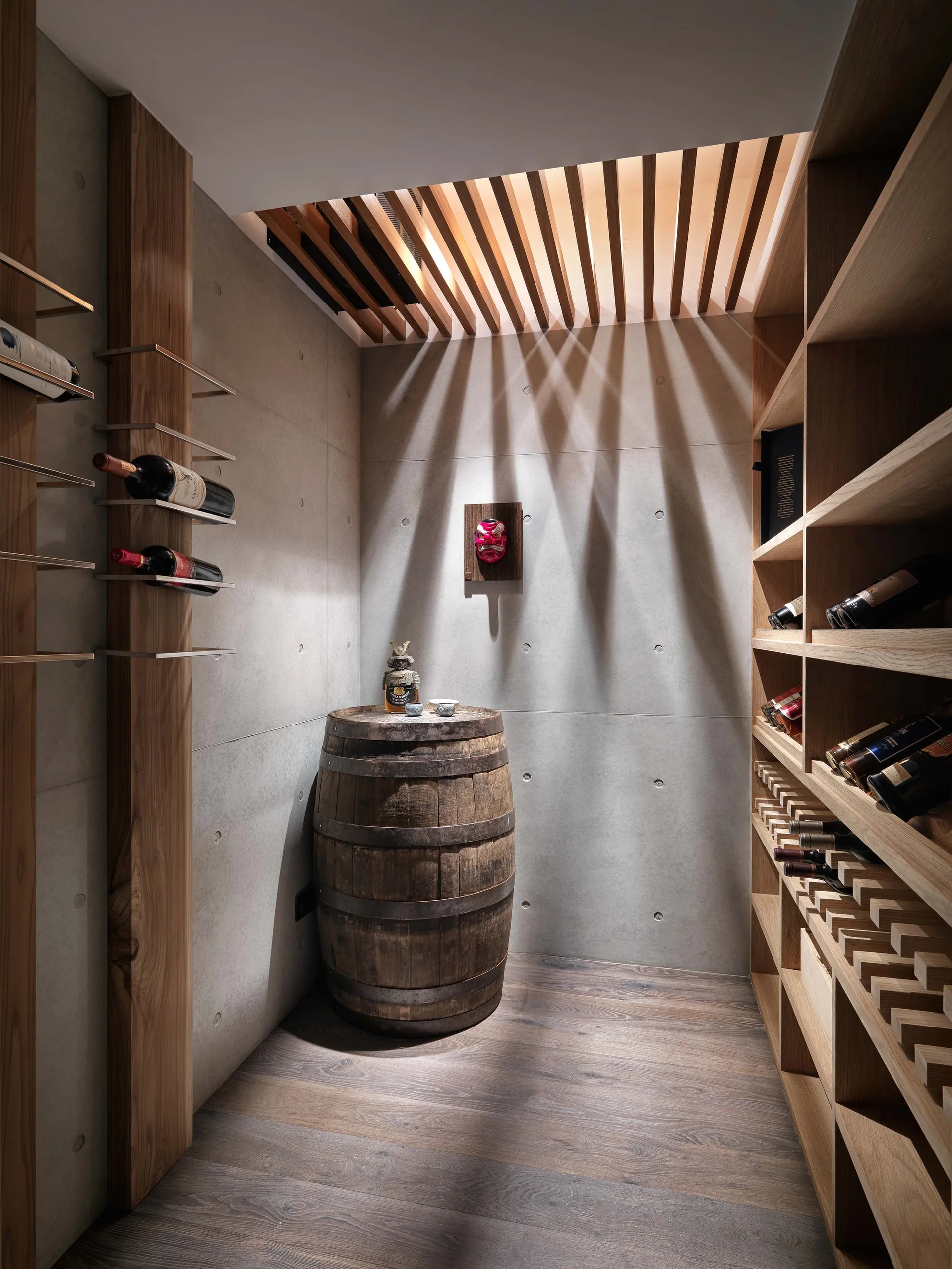 A minimalist wine cellar with exposed concrete walls, custom wooden shelving, and an old wine barrel under a slatted ceiling that casts dramatic shadows.