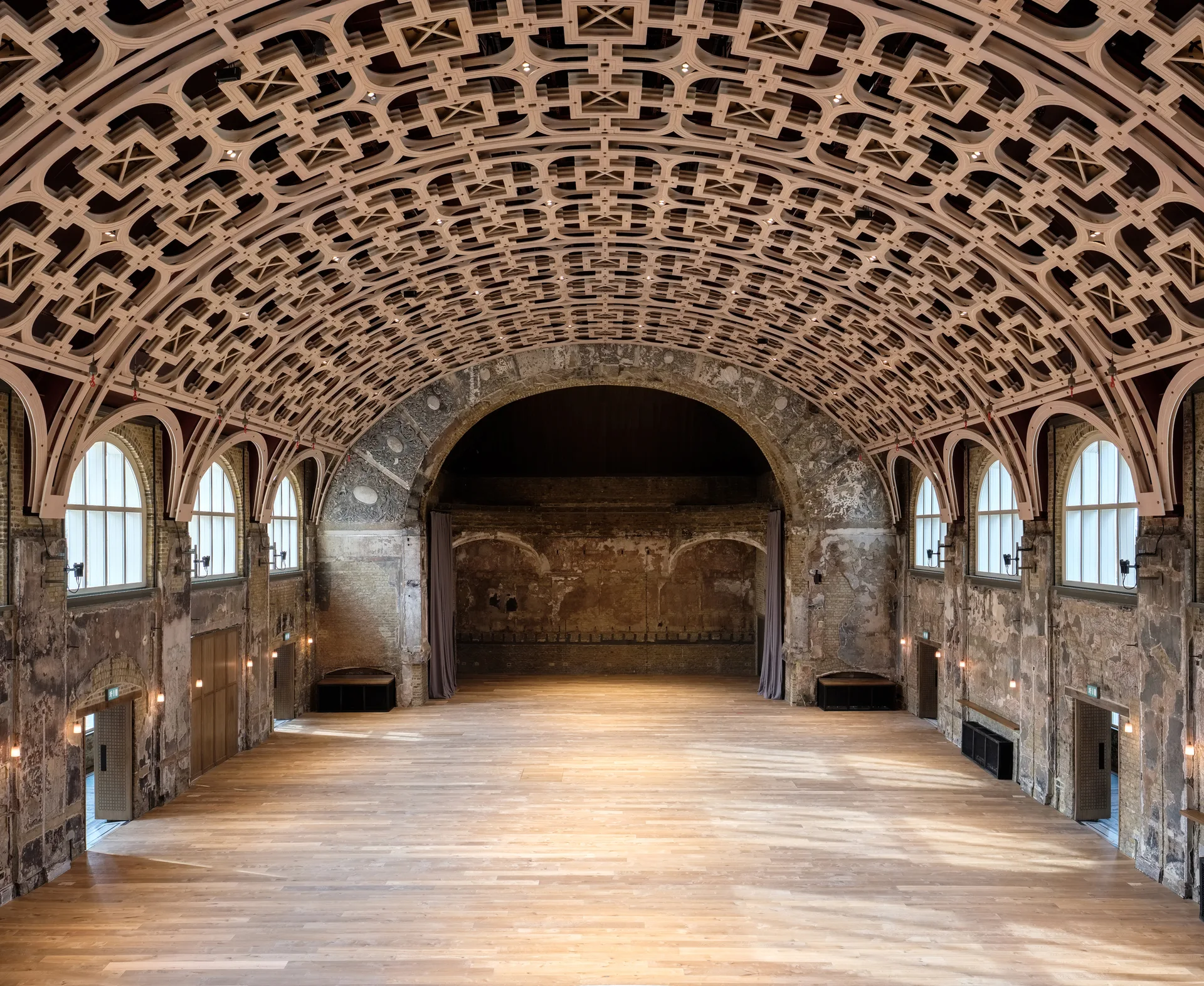 A spacious historic hall features an ornate geometric vaulted ceiling, distressed plaster walls with arched windows, and a light wood plank floor.