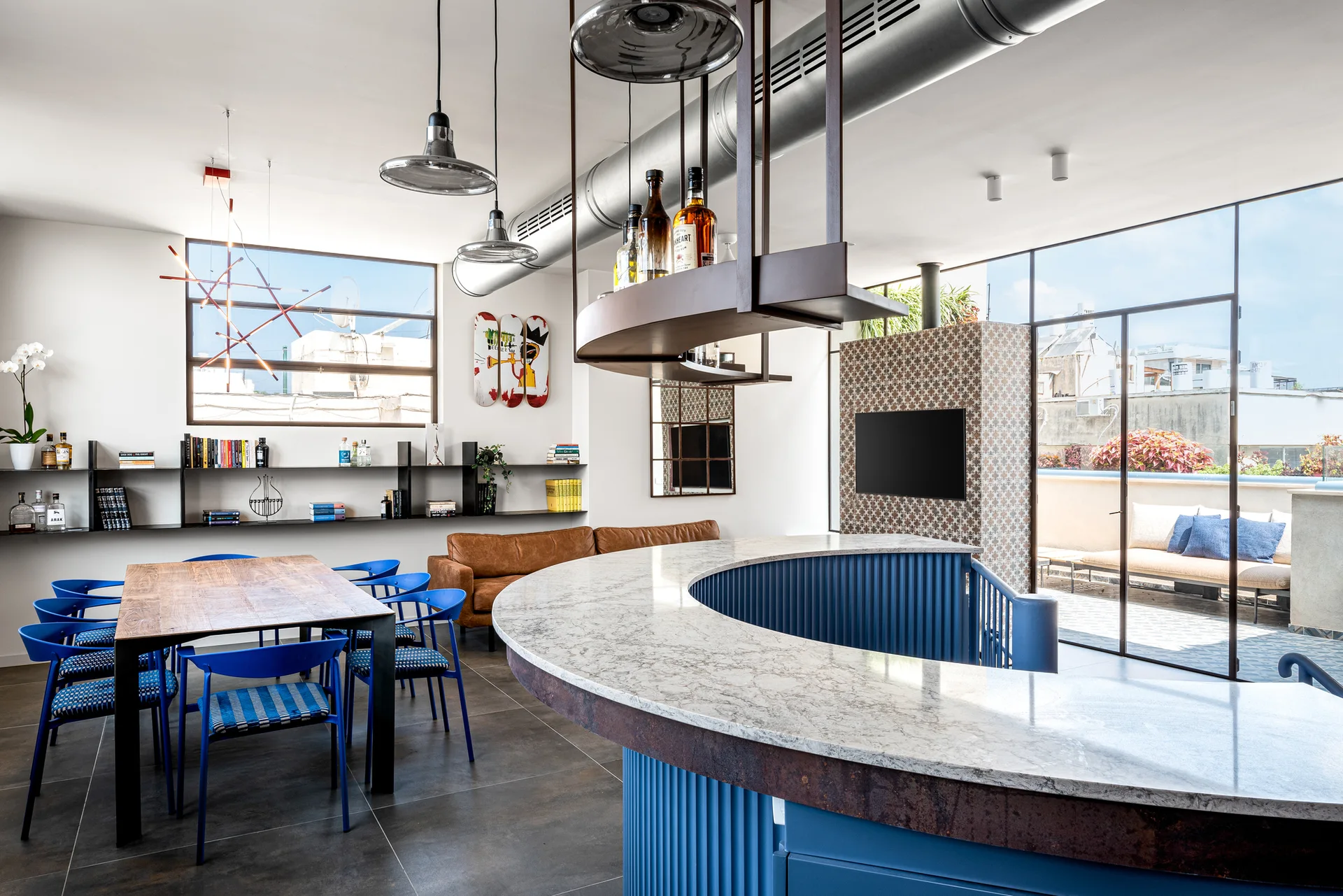 A modern industrial open living area featuring a large curved blue bar, a wooden dining table with blue chairs, and an exposed silver ceiling duct.