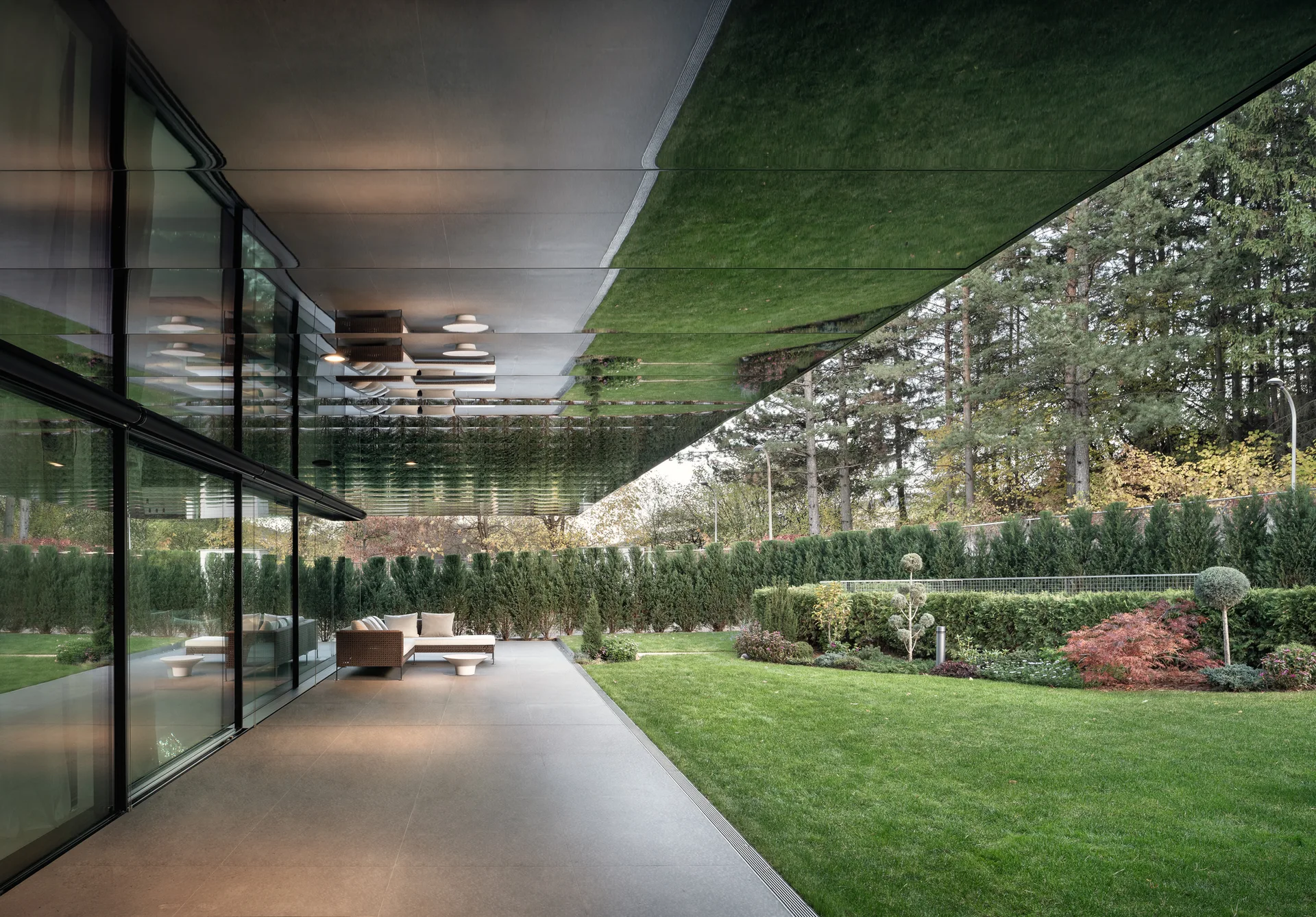 A modern covered patio with a wicker sectional sofa beneath a reflective cantilevered roof overlooking a manicured lawn.