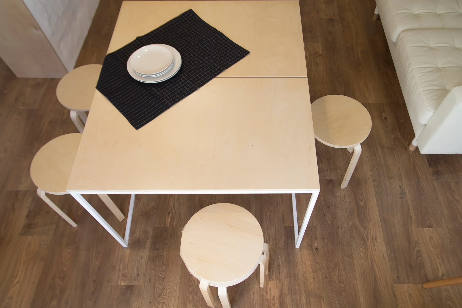 An overhead view of a light wood dining table with four matching round stools and a black checkered placemat on wood flooring.