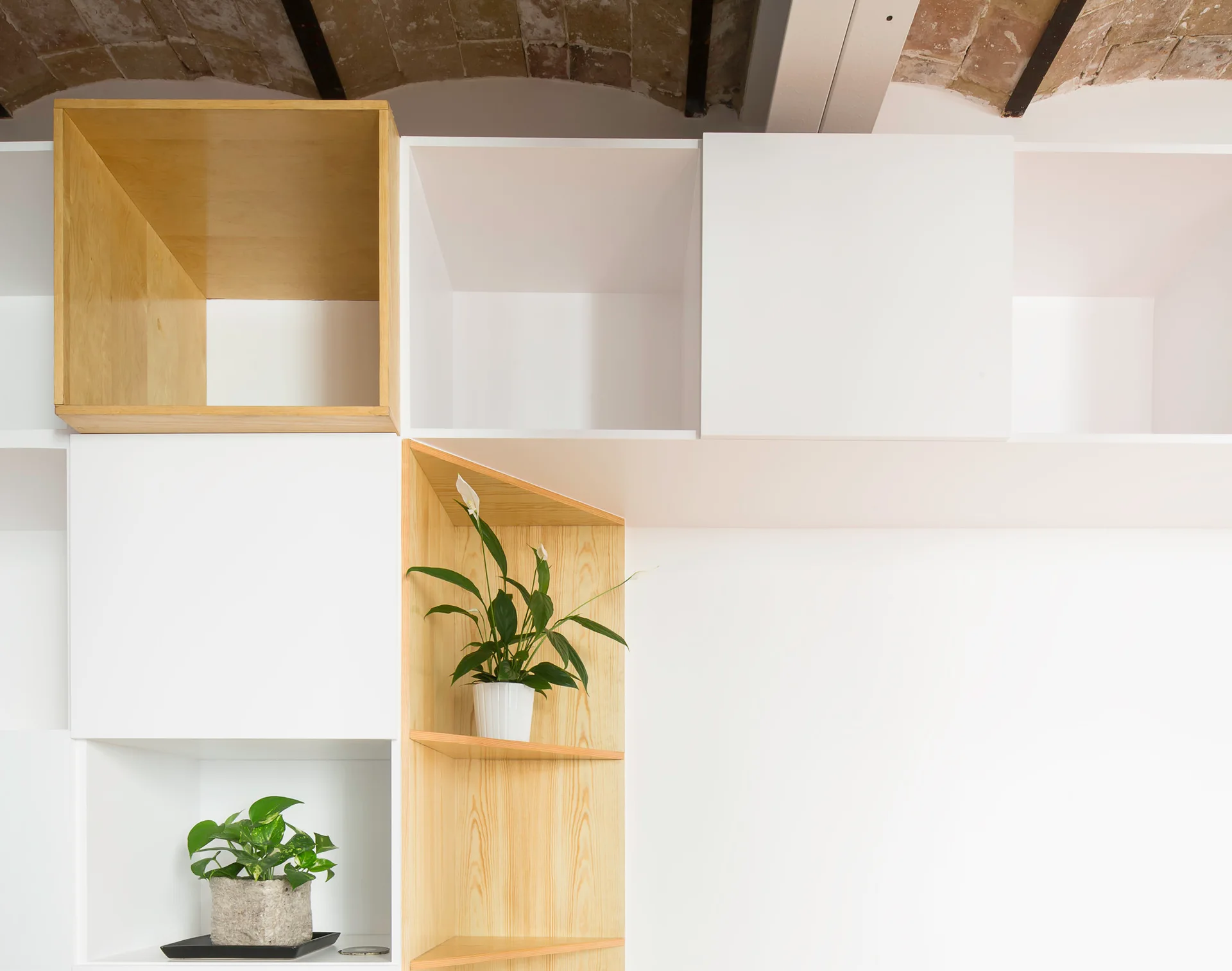 A modern shelving unit with white and natural wood compartments holds small potted plants beneath an exposed brick vaulted ceiling.
