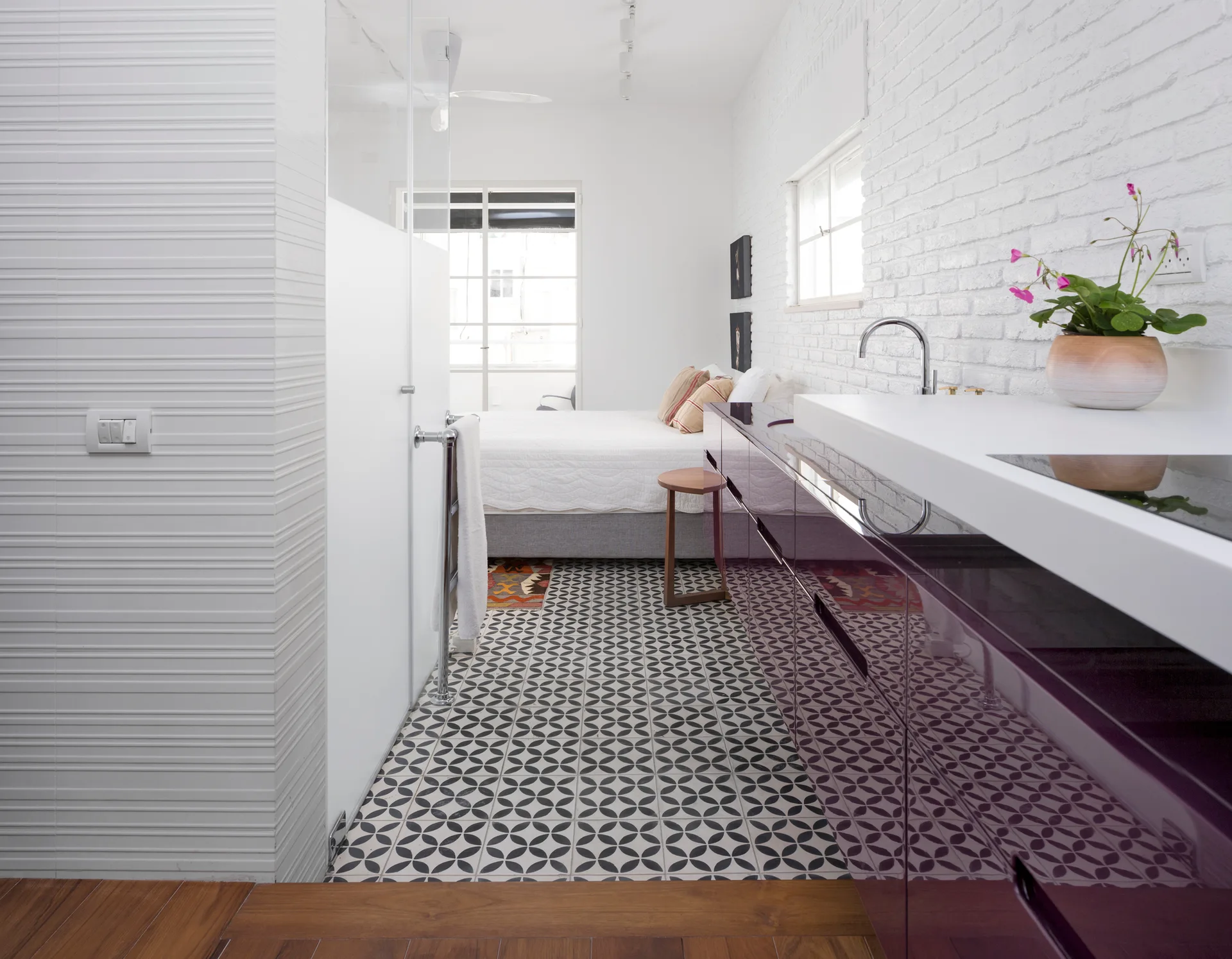 A modern bedroom suite featuring a glossy purple vanity, a white painted brick wall, and geometric patterned floor tiles leading to a bed.