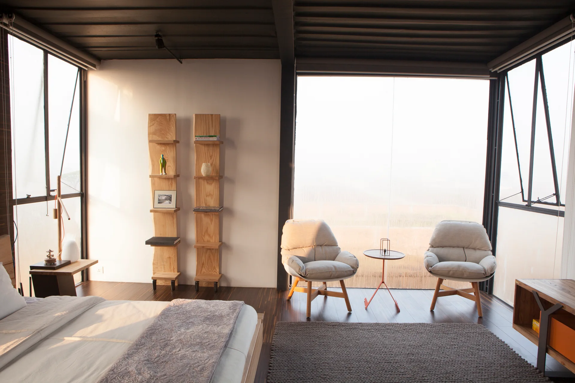 Modern bedroom with a bed, two grey armchairs by large windows, and vertical wooden shelves against a white wall under a dark beamed ceiling.