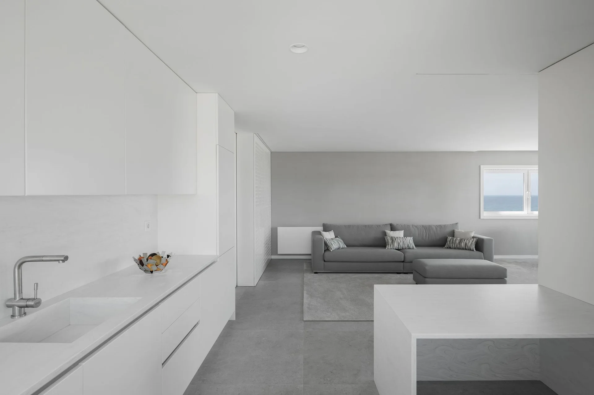 An open-plan kitchen and living room featuring seamless white cabinetry, an integrated sink, and a grey sofa on a large area rug.